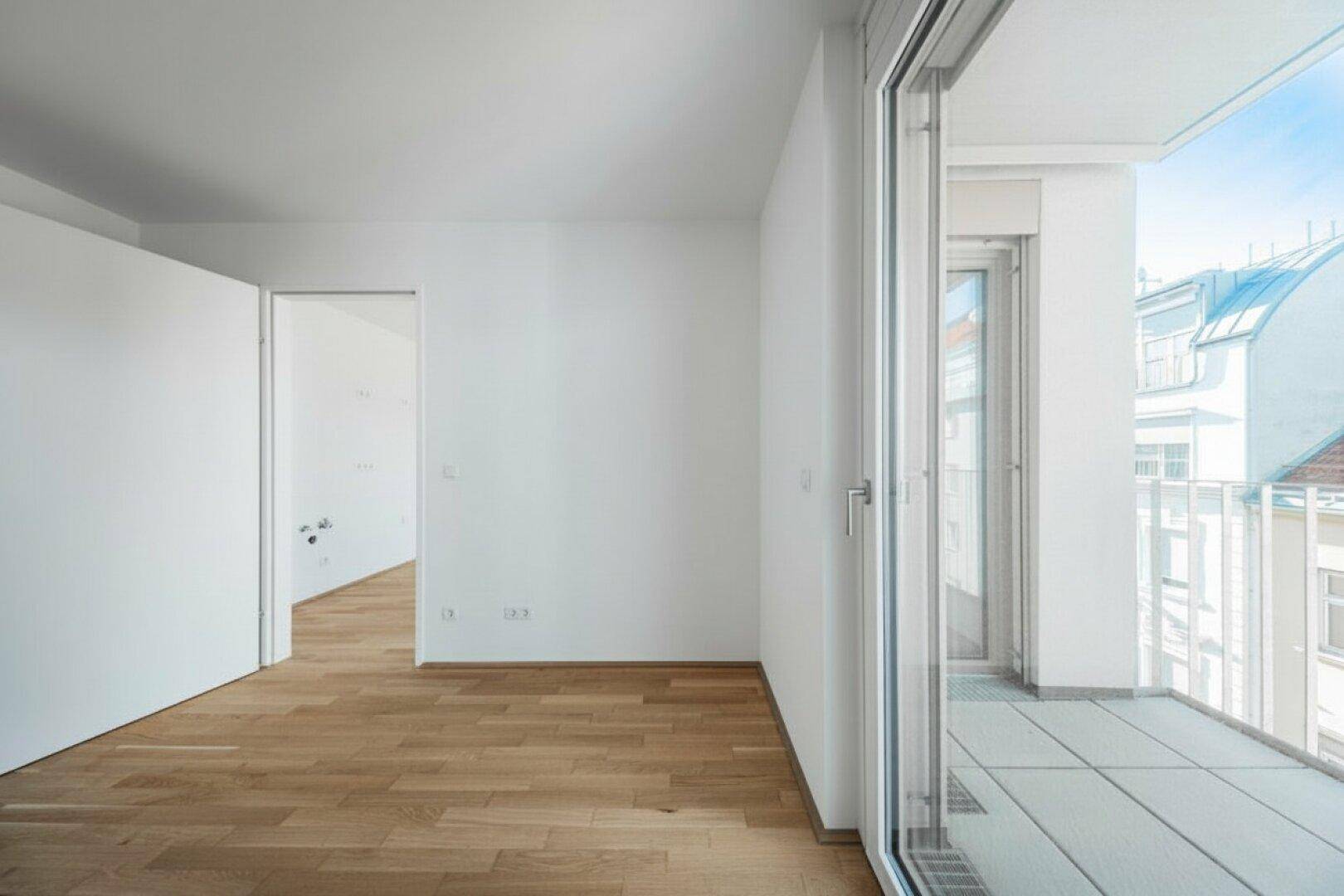 Geräumiger, heller Raum mit Holzboden und breitem Zugang zu einem sonnigen Balkon.