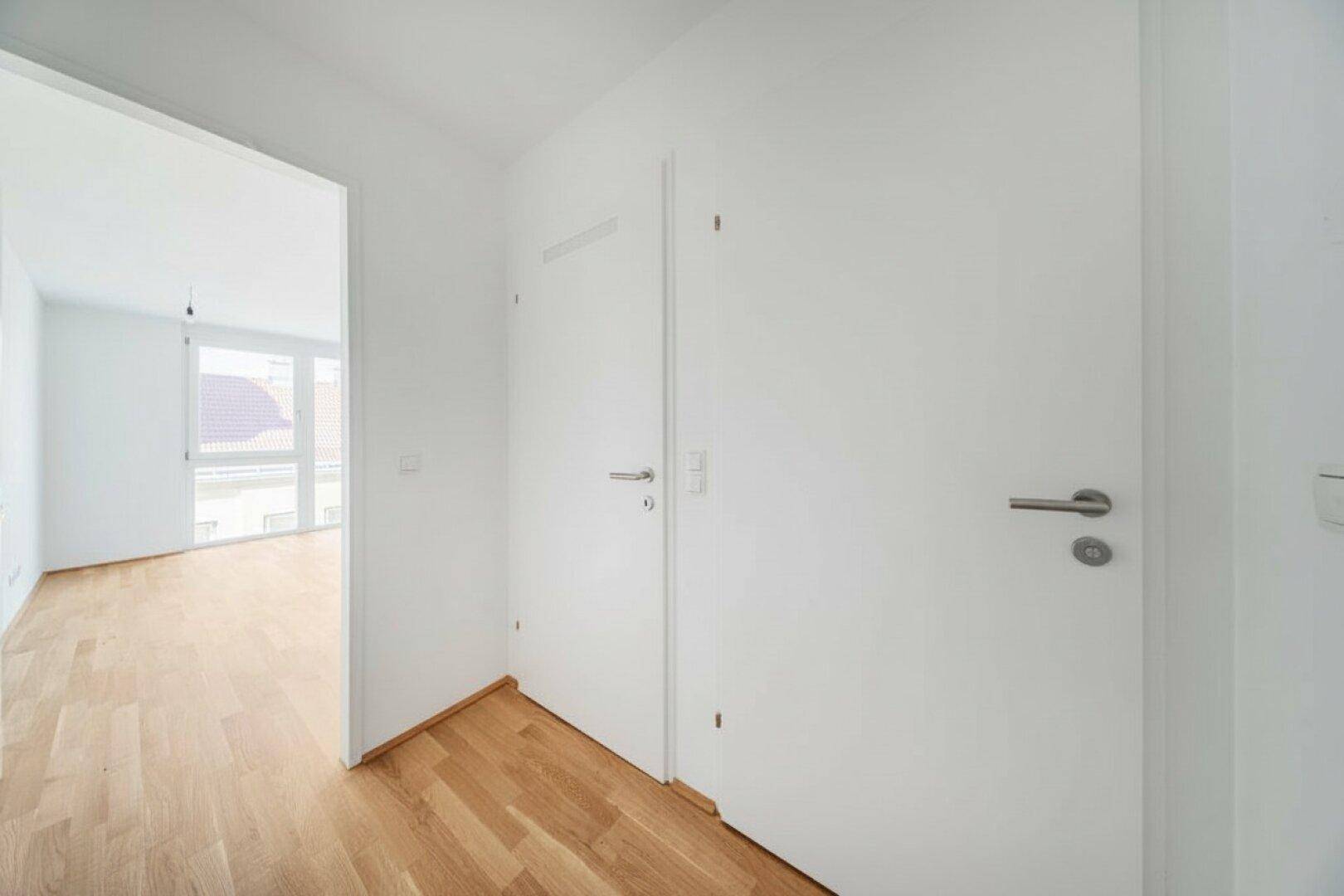 Schlichter Flur mit Holzboden und weißen Wänden, der zu weiteren Räumen führt.