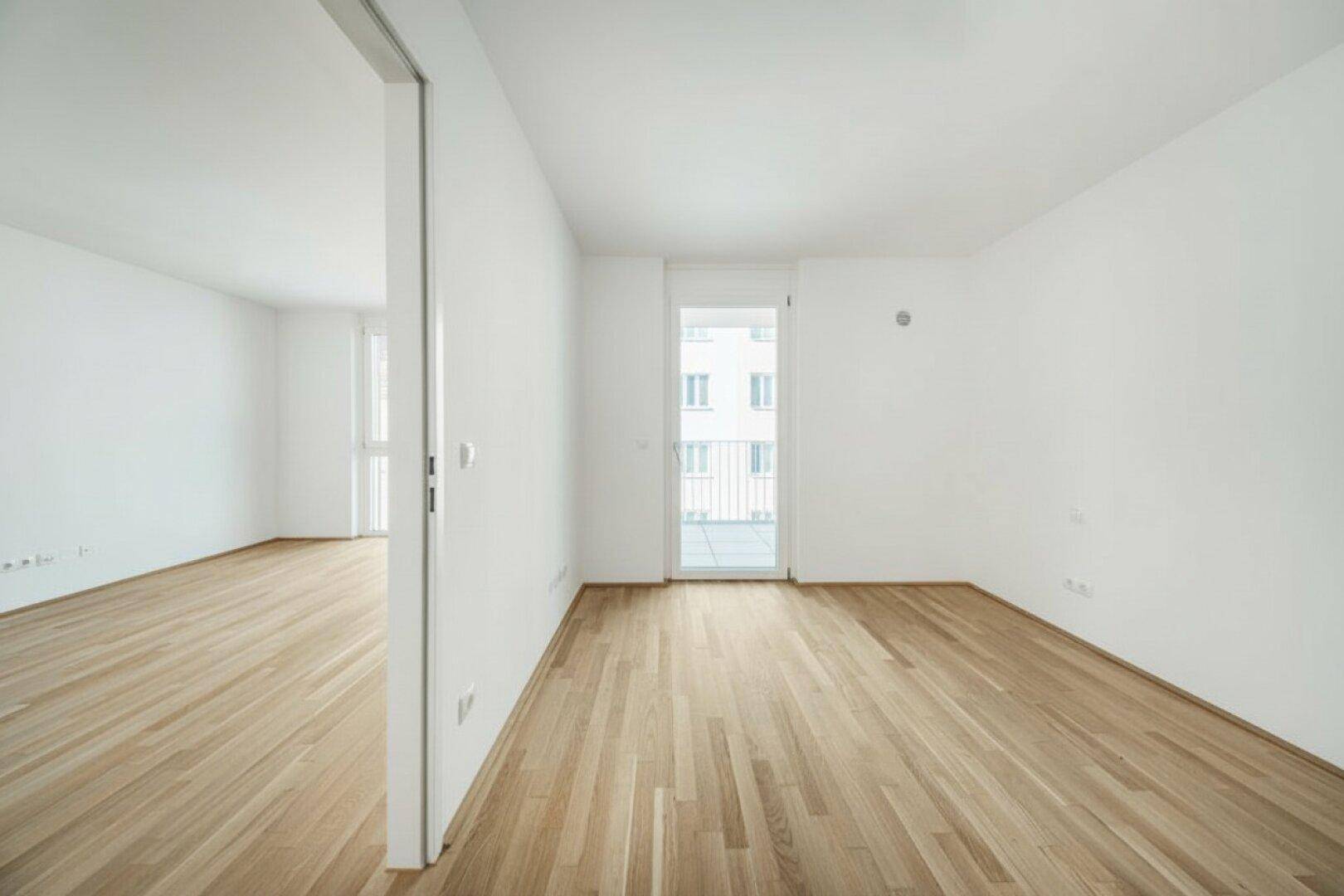 Helles, leeres Zimmer mit Parkettboden und Zugang zu einem Balkon sowie einem weiteren Raum.