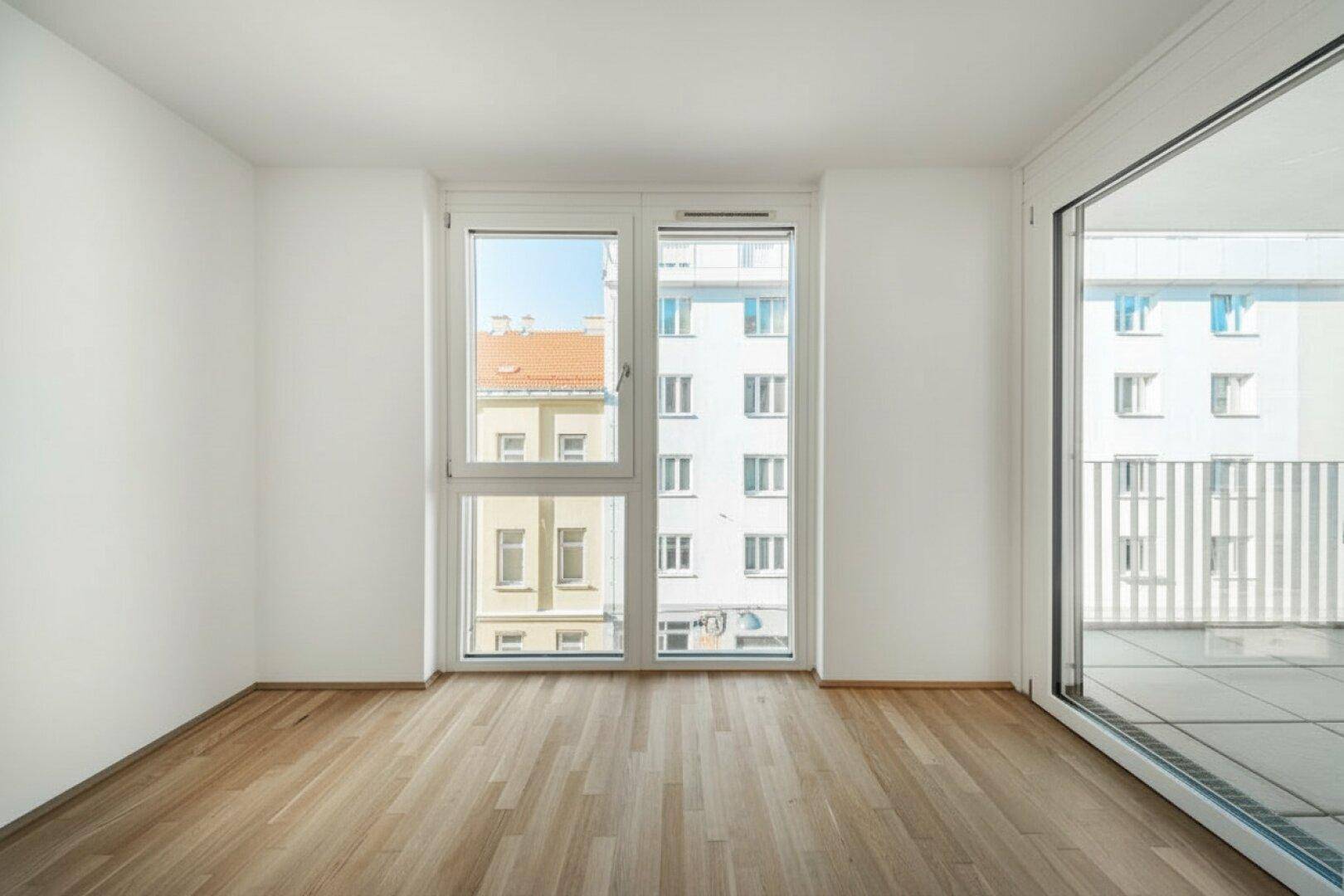 Helles, leeres Zimmer mit Parkettboden, Fenstern und Zugang zu einem Balkon.
