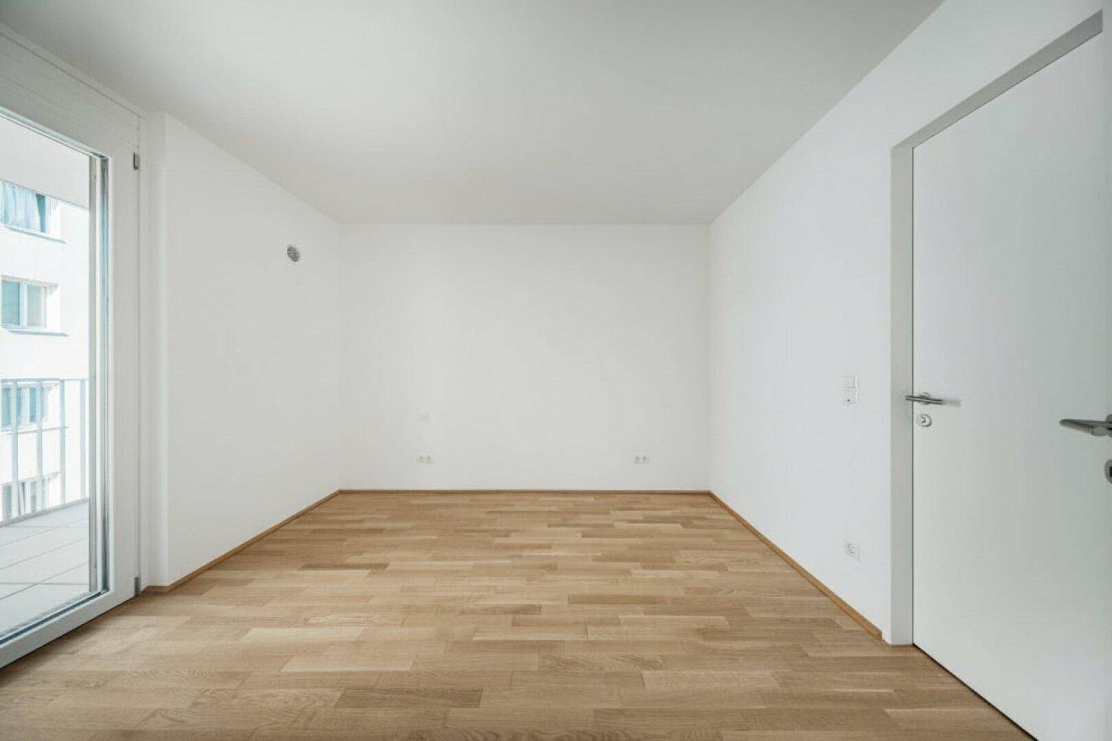 Leerer Raum mit Parkettboden, weißer Tür und Zugang zum Außenbereich.