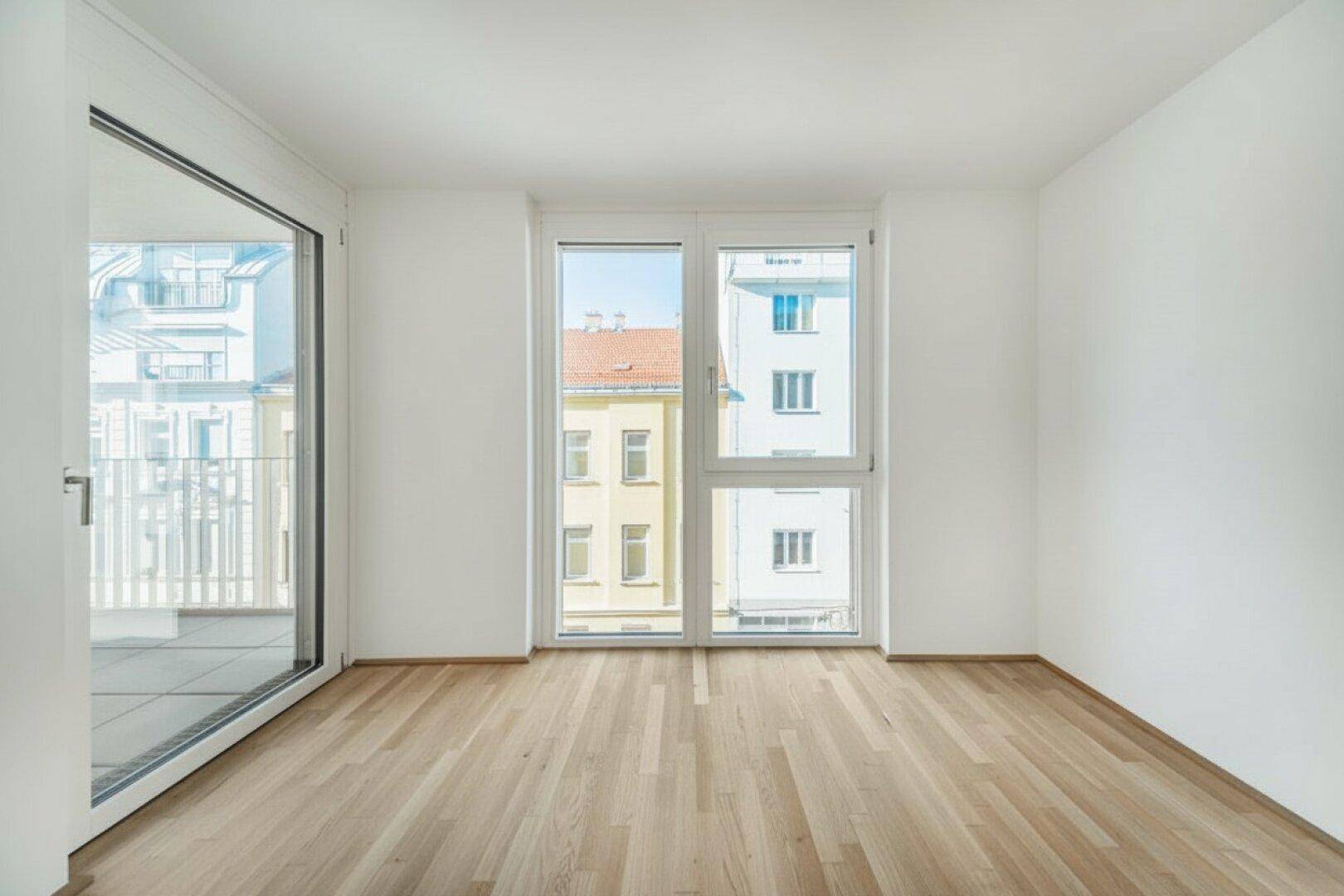 Heller Raum mit Parkettboden und großen Fenstern, die einen Blick auf die umliegenden Gebäude bieten.
