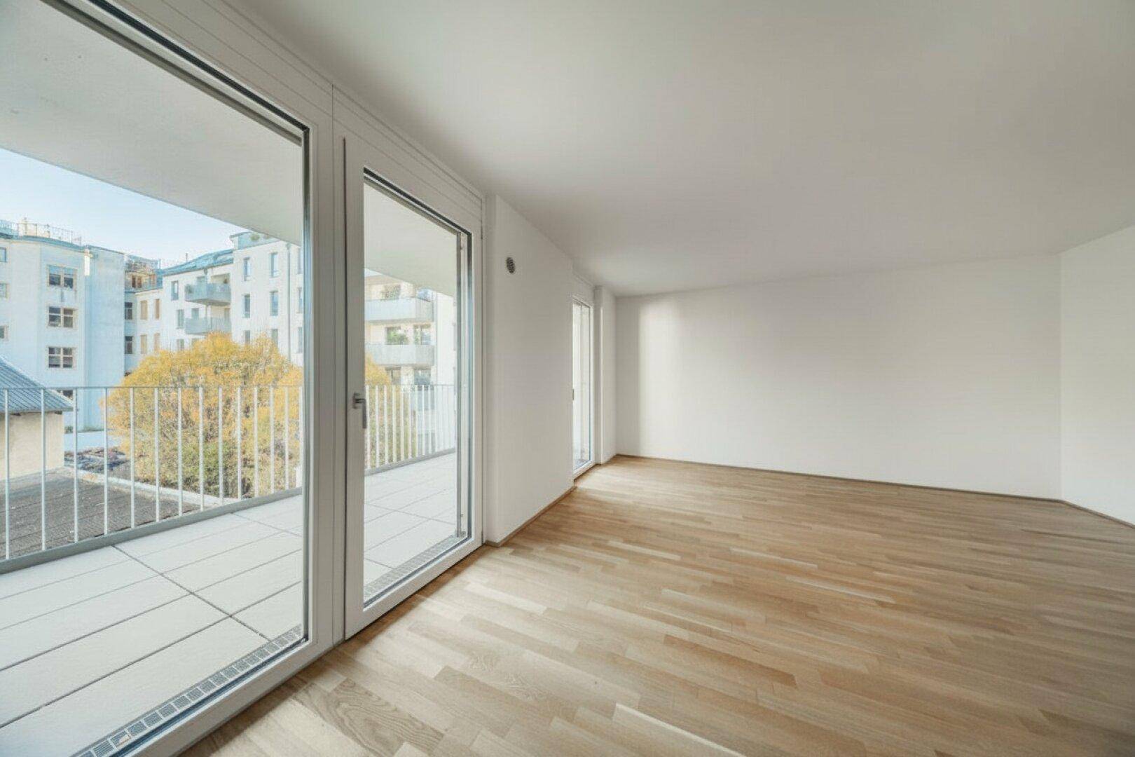 Geräumiger, leerer Raum mit Holzboden und Zugang zu einem sonnigen Balkon.