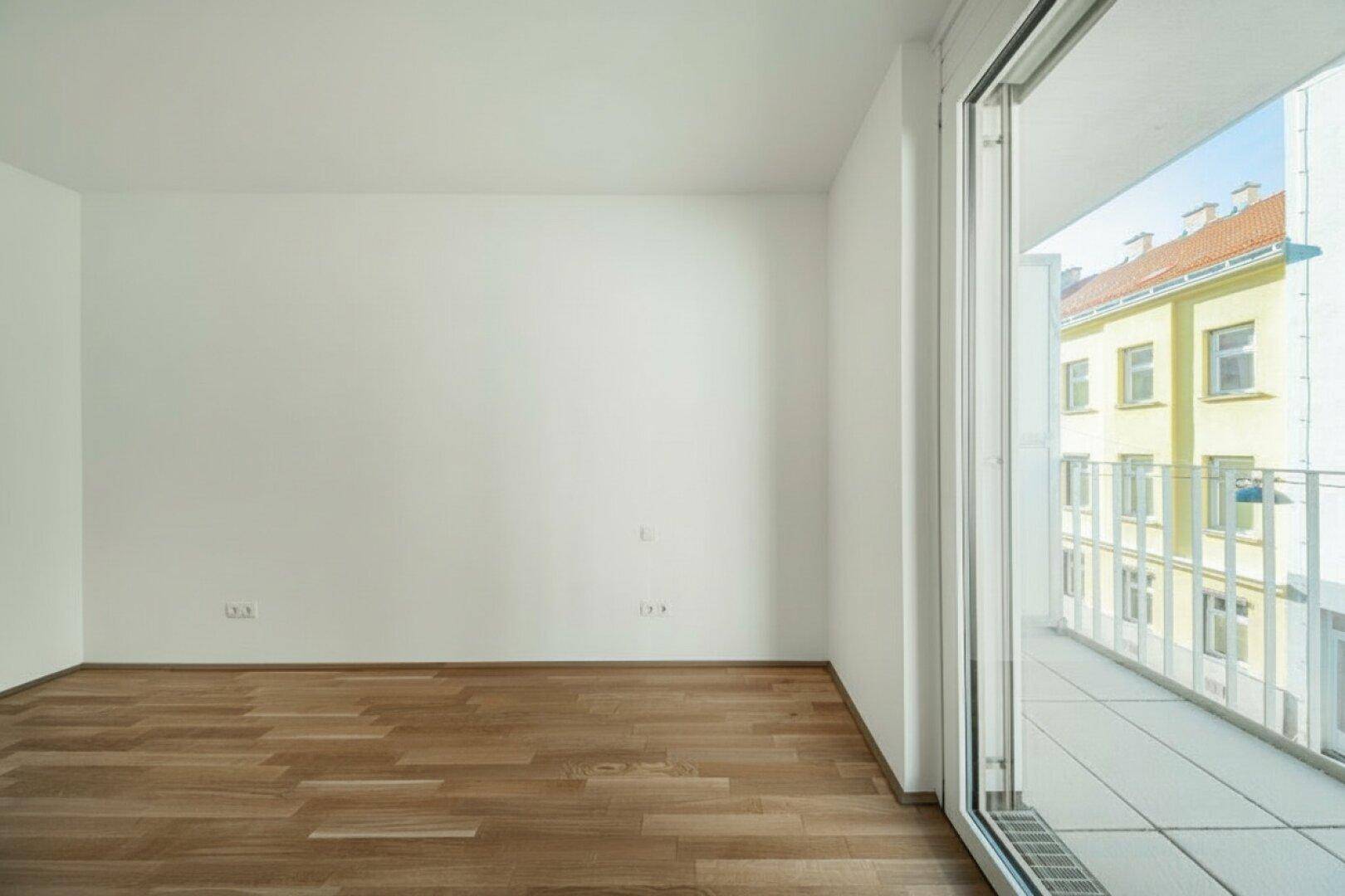 Heller Raum mit Holzboden und großem Schiebefenster zum Balkon, Blick auf Nachbargebäude.