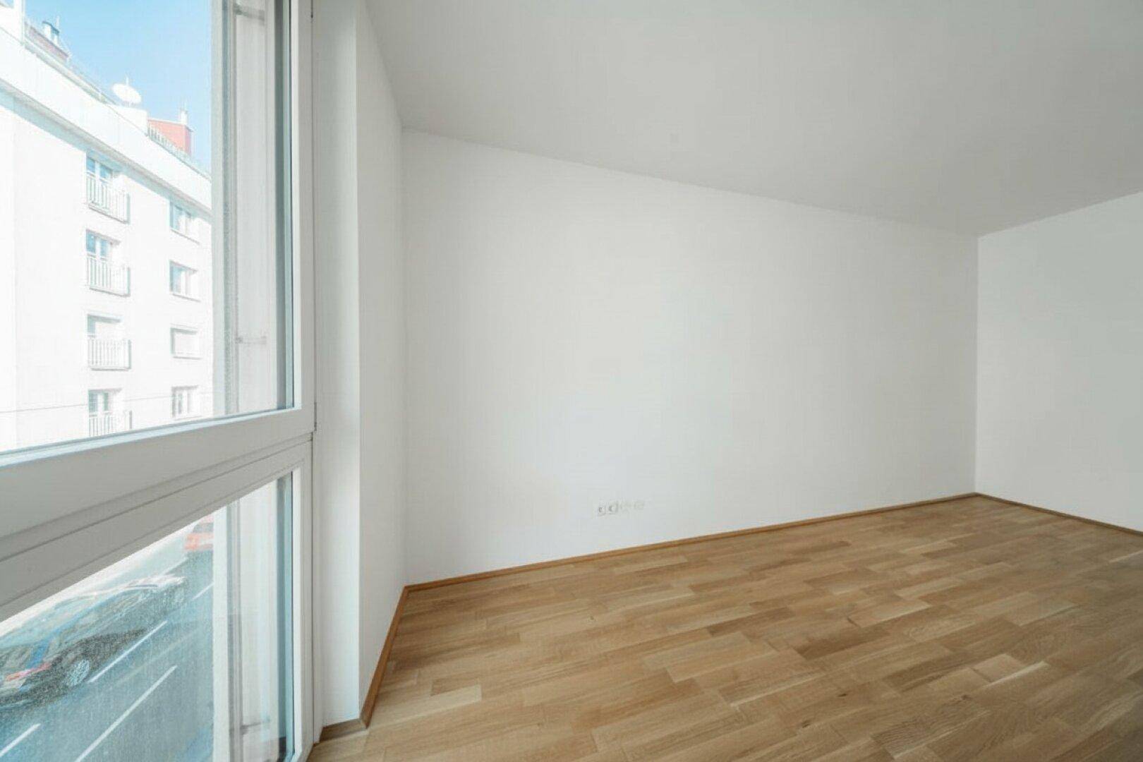Leerer Raum mit Holzboden und großem Fenster, das viel Tageslicht hereinlässt.