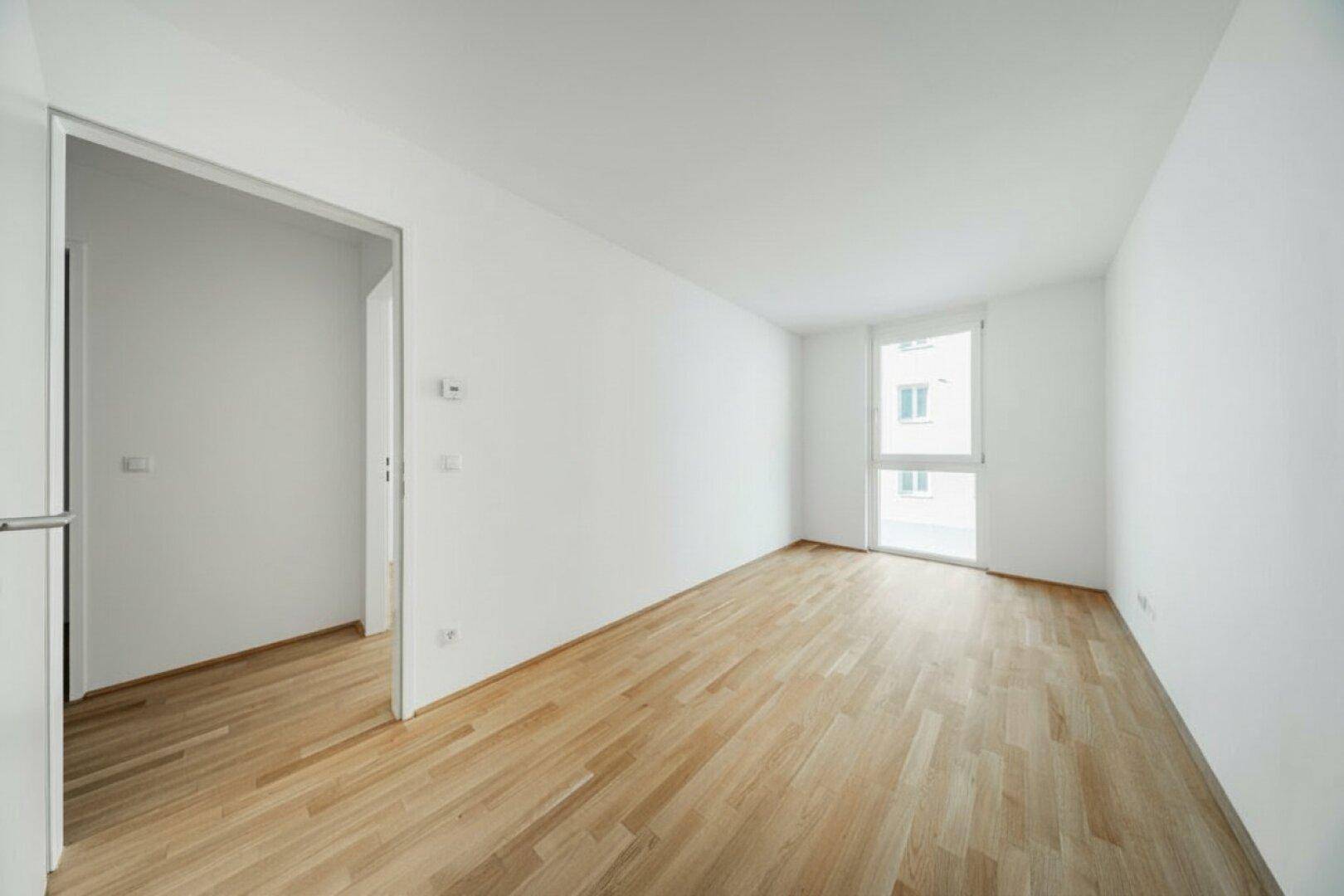 Heller Raum mit Holzboden und großem Fenster, offen zu einem weiteren Bereich.