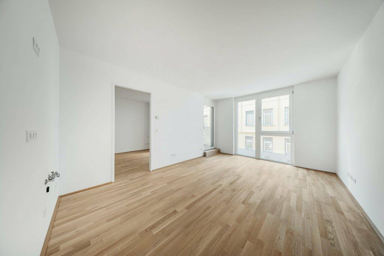 Geräumiger, heller Raum mit Holzboden, großen Fenstern und Balkonzugang.