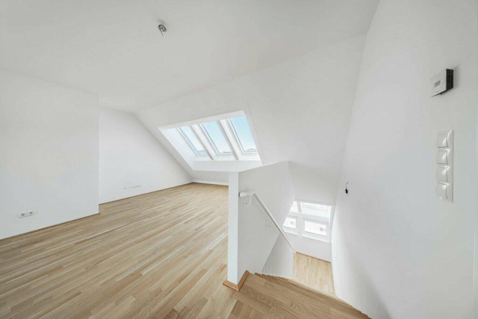 Heller, leerer Raum im Dachgeschoss mit Holzboden, weißen Wänden und mehreren Dachfenstern.