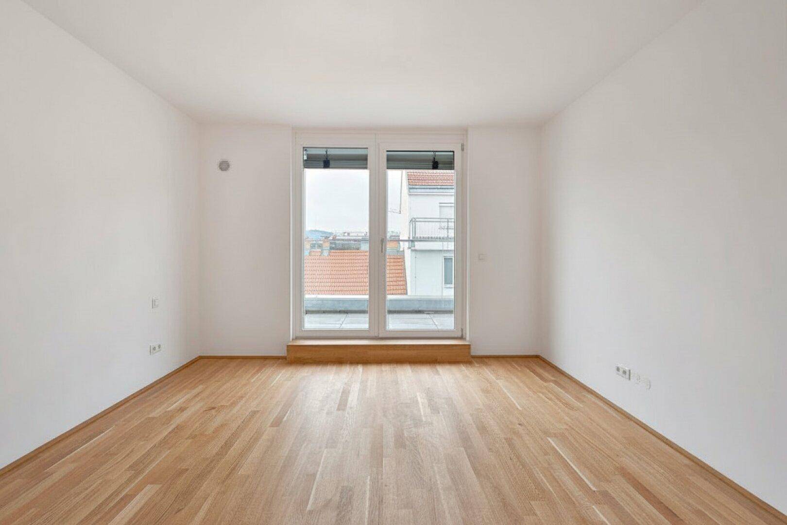 Heller, leerer Raum mit Holzboden und großem Fenster, das viel Tageslicht hereinlässt.