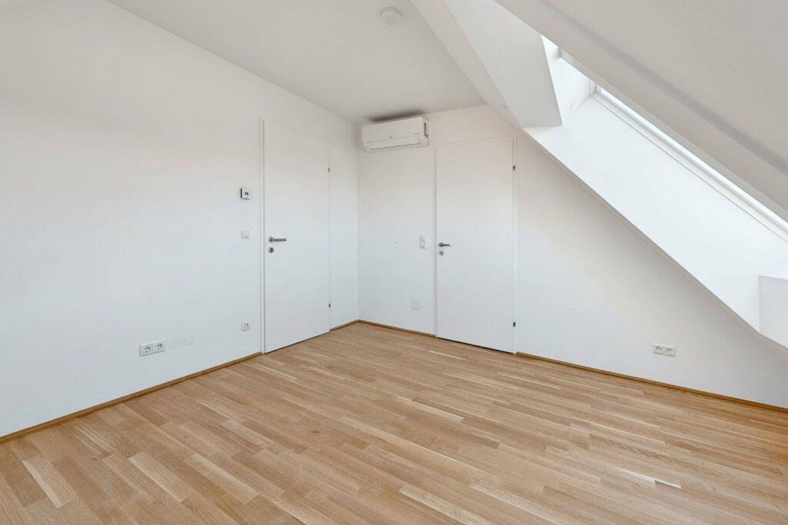 Heller Dachgeschossraum mit Holzboden, zwei Türen und Klimaanlage für angenehmes Raumklima.