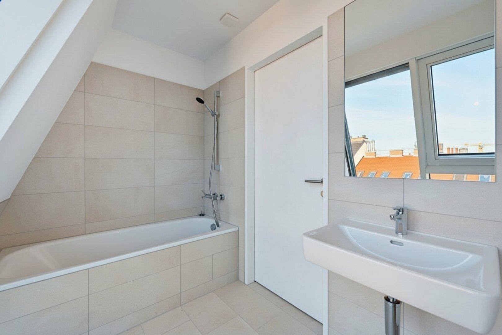 Modernes Badezimmer mit Badewanne, Waschbecken und hellen Fliesen, unter einer Dachschräge gelegen.