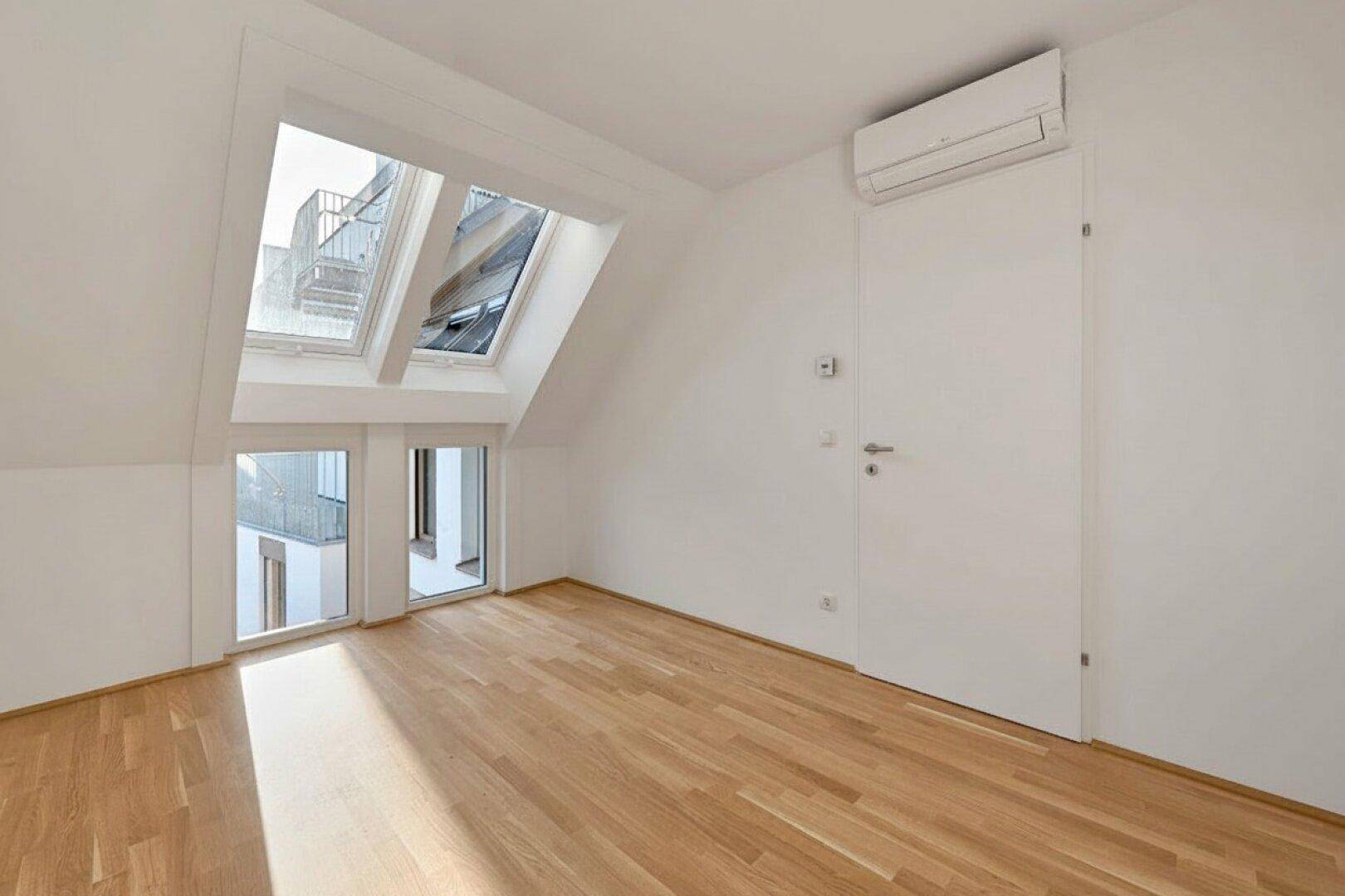 Helles Dachgeschosszimmer mit Parkettboden, Dachfenstern und Blick auf die umliegenden Gebäude.