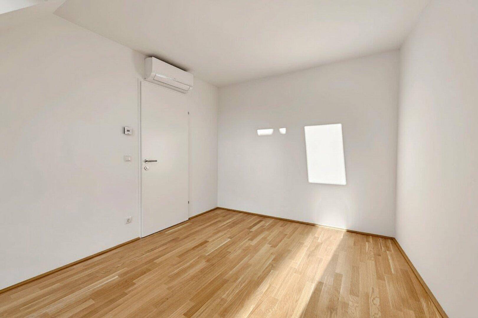 Helles Zimmer mit Parkettboden, Klimaanlage und natürlichem Lichteinfall durch ein kleines Fenster.