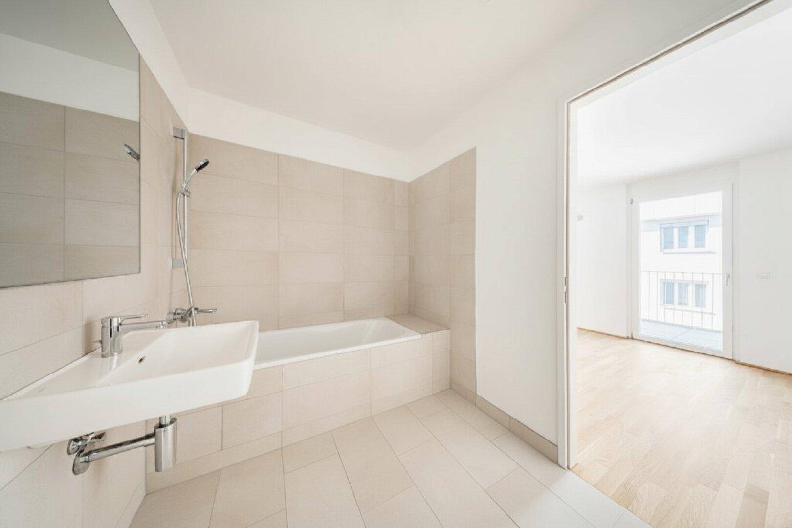 Helles Badezimmer mit Badewanne, Waschbecken und Fliesenboden, mit Zugang zu einem Zimmer.