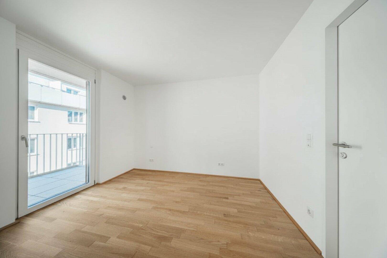 Leeres Zimmer mit Holzboden und Balkontür, bietet direkten Zugang zum Außenbereich.