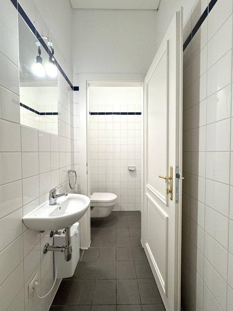 Schlichtes Badezimmer mit weißen Wandfliesen, dunklen Bodenfliesen und einem kleinen Waschbecken und Toilette.