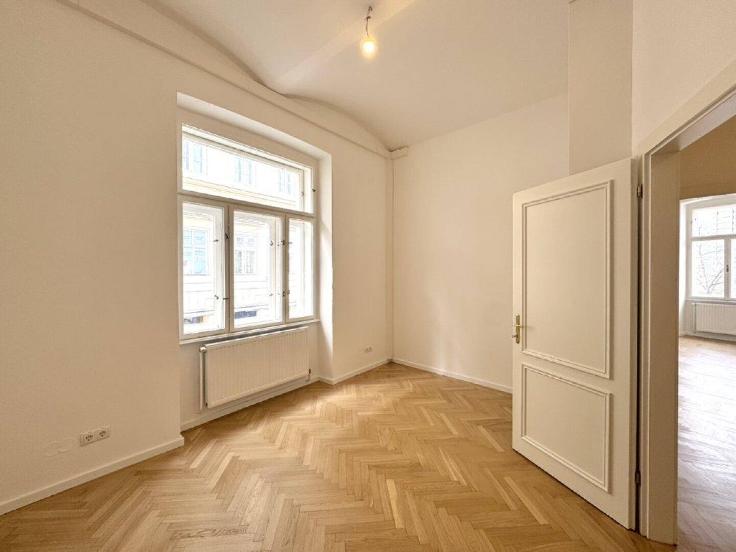 Helles Zimmer mit Fischgrätparkett, weißen Wänden und einem großen Fenster, das viel Licht spendet und eine offene Tür.