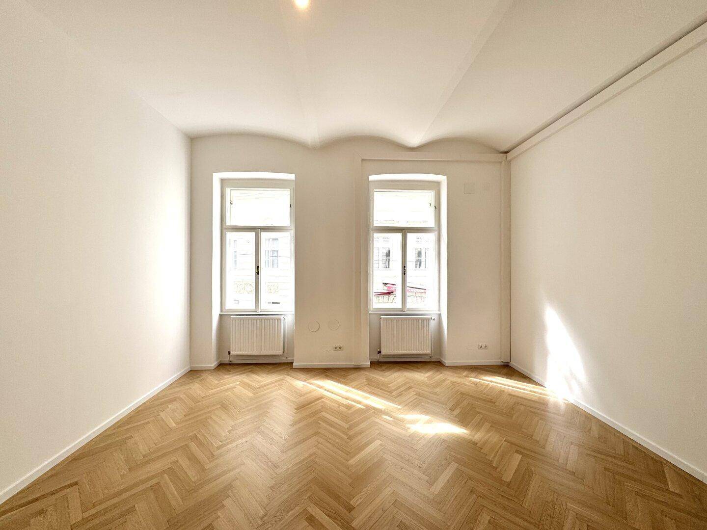 Heller, leerer Raum mit Fischgrätparkett, weißen Wänden und zwei großen Fenstern, die den Raum mit Sonne füllen.