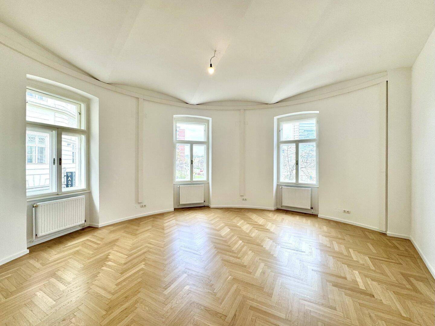 Heller, leerer Raum mit Fischgrätparkett, weißen Wänden und drei großen Fenstern, die viel Tageslicht hereinlassen.