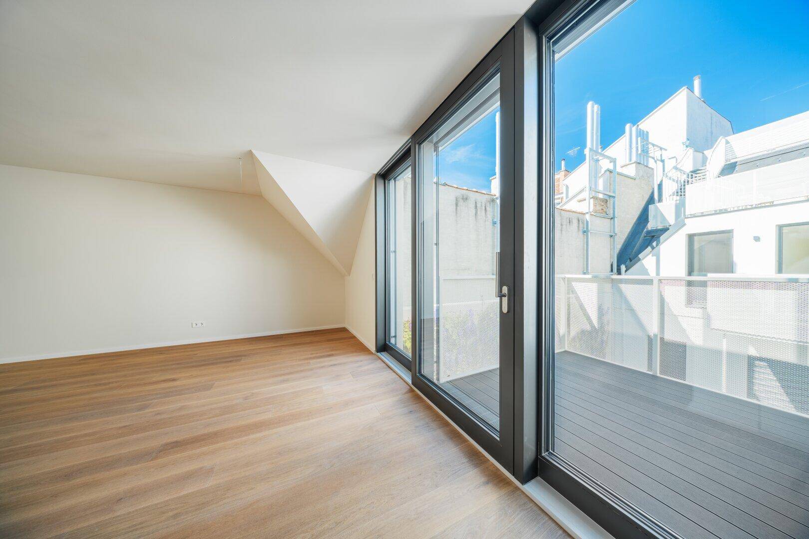 Lichtdurchfluteter Raum mit Parkettboden und Zugang zu einer Terrasse, umgeben von modernen Gebäuden.