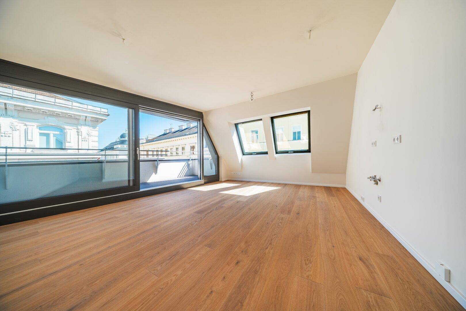 Großer, heller Raum mit Holzboden und breiter Fensterfront, die Zugang zu einem Balkon bietet.