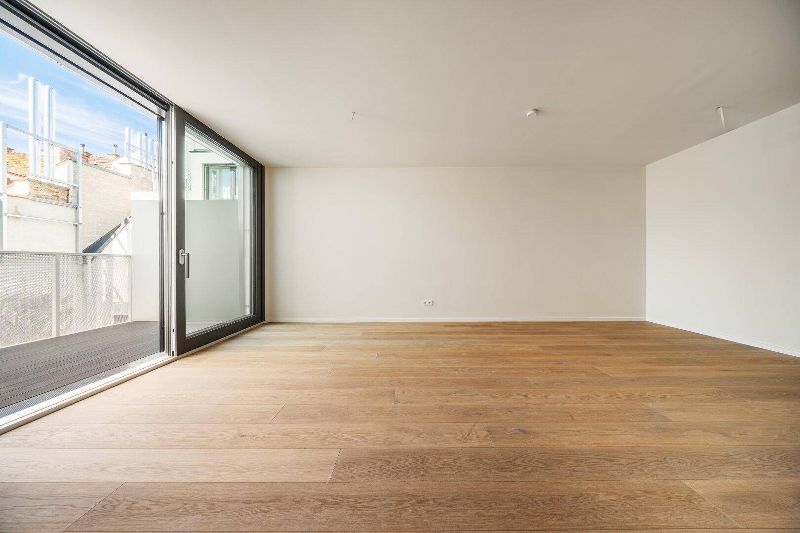 Geräumiger, heller Raum mit Holzboden und großem Schiebefenster zum Balkon, unmöbliert.