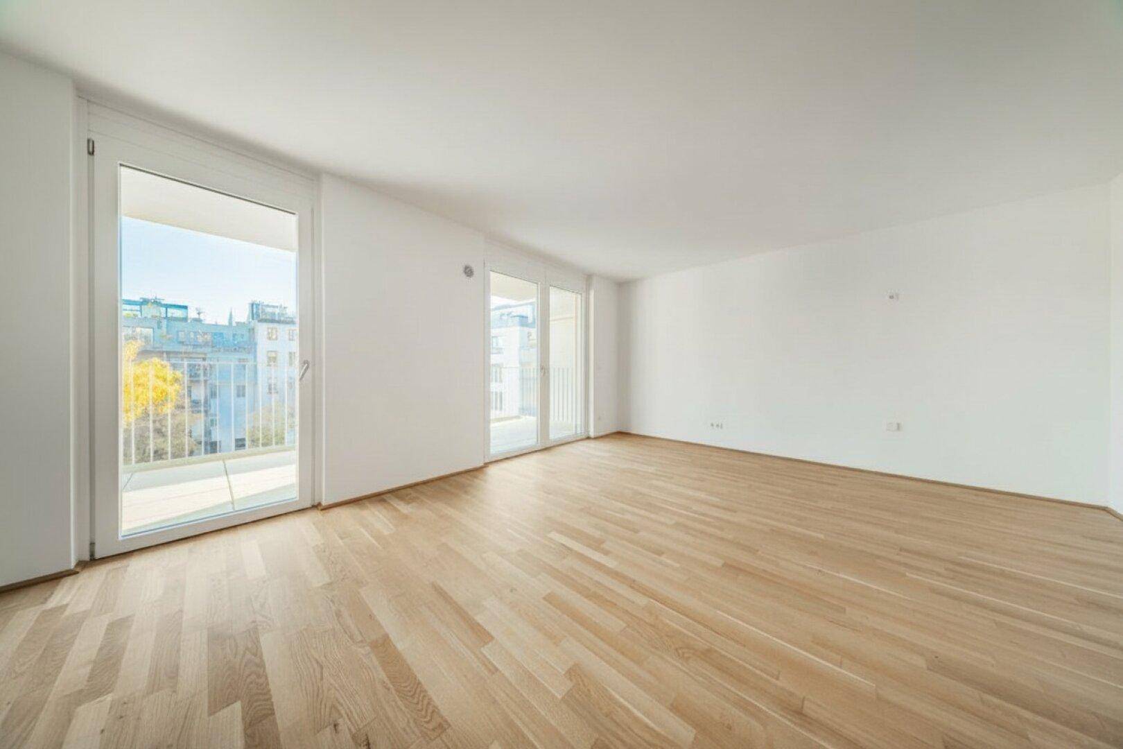 Geräumiges, leeres Wohnzimmer mit Parkettboden und mehreren Balkonzugängen, sehr hell.
