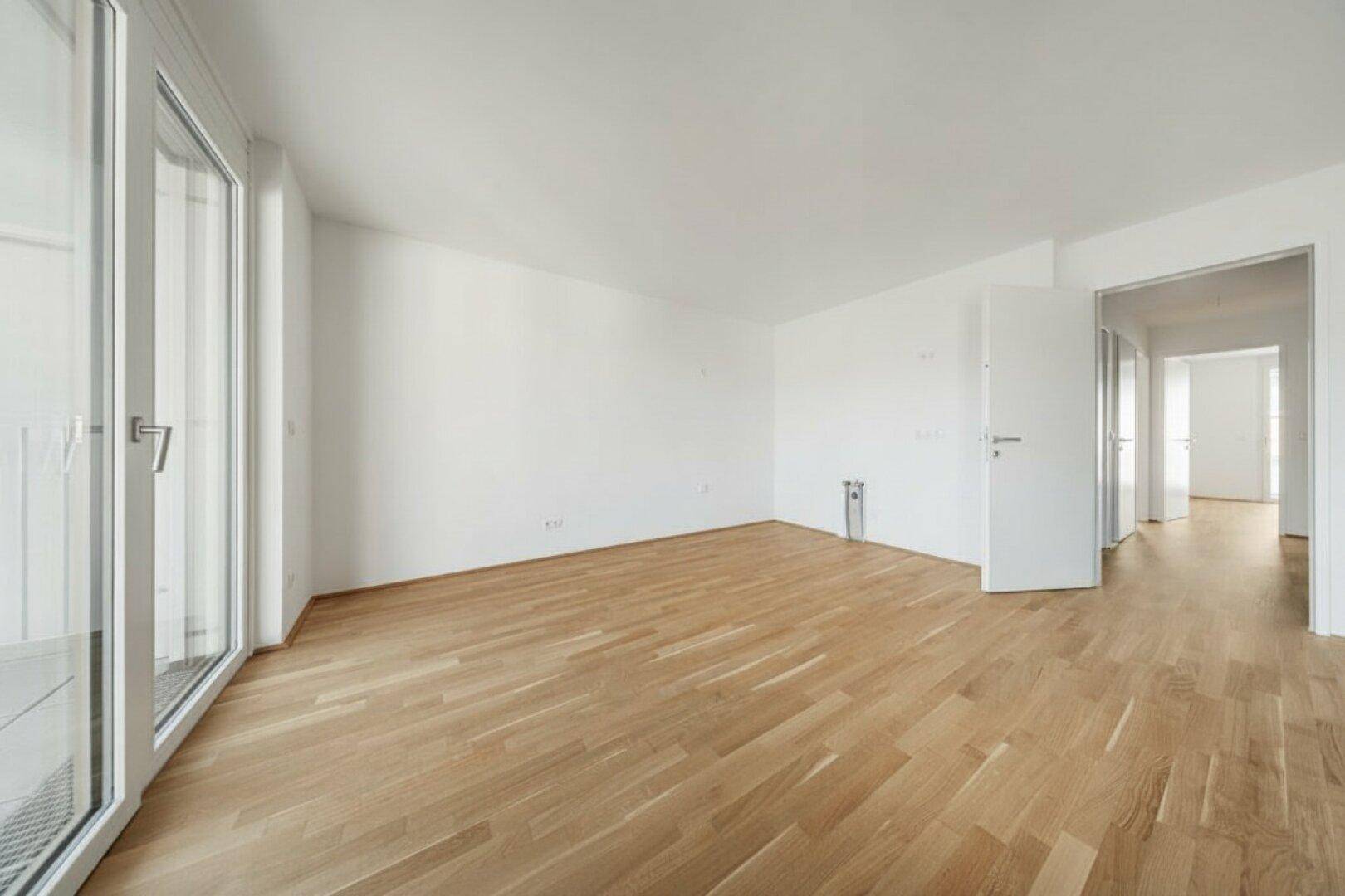 Geräumiges, leeres Wohnzimmer mit Parkettboden und Zugang zu einem Balkon, sehr hell.