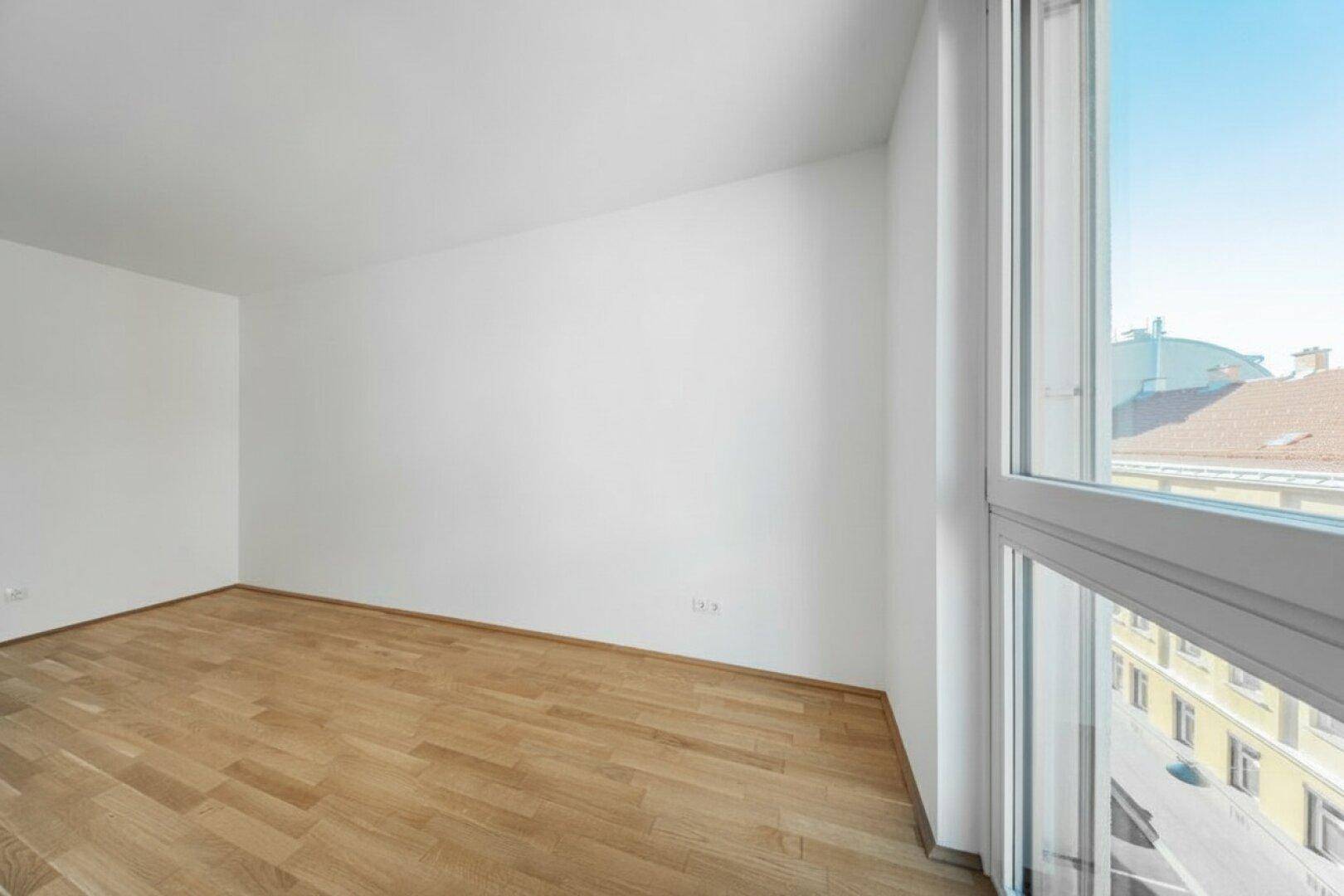 Heller, leerer Raum mit Parkettboden und großem Fenster, bietet viel Tageslicht.