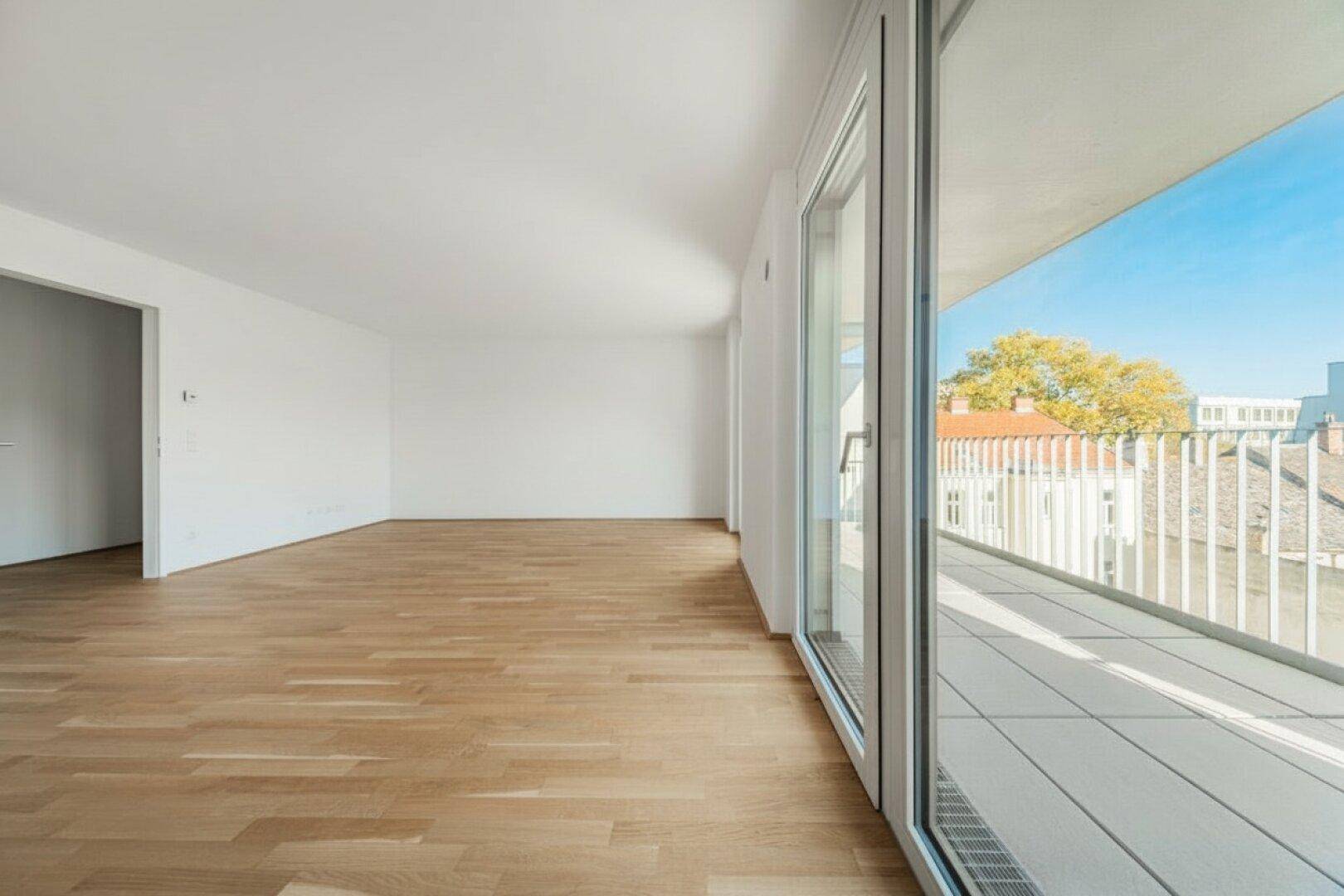 Helles, leeres Zimmer mit Parkettboden und Zugang zu einem breiten Balkon mit Stadtblick.