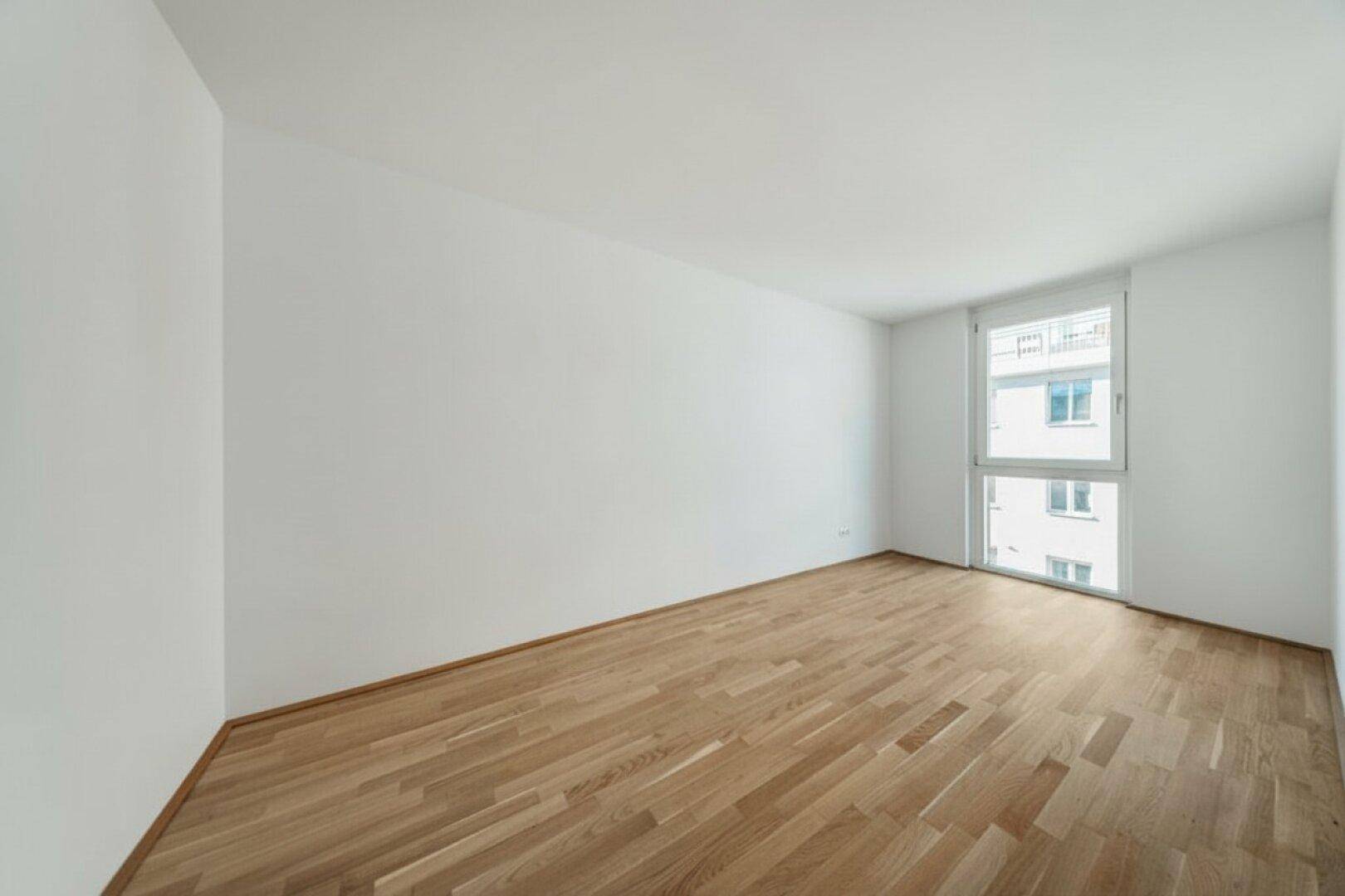 Leeres Schlafzimmer mit hellem Parkettboden und einem großen Fenster, das viel Tageslicht hereinlässt.