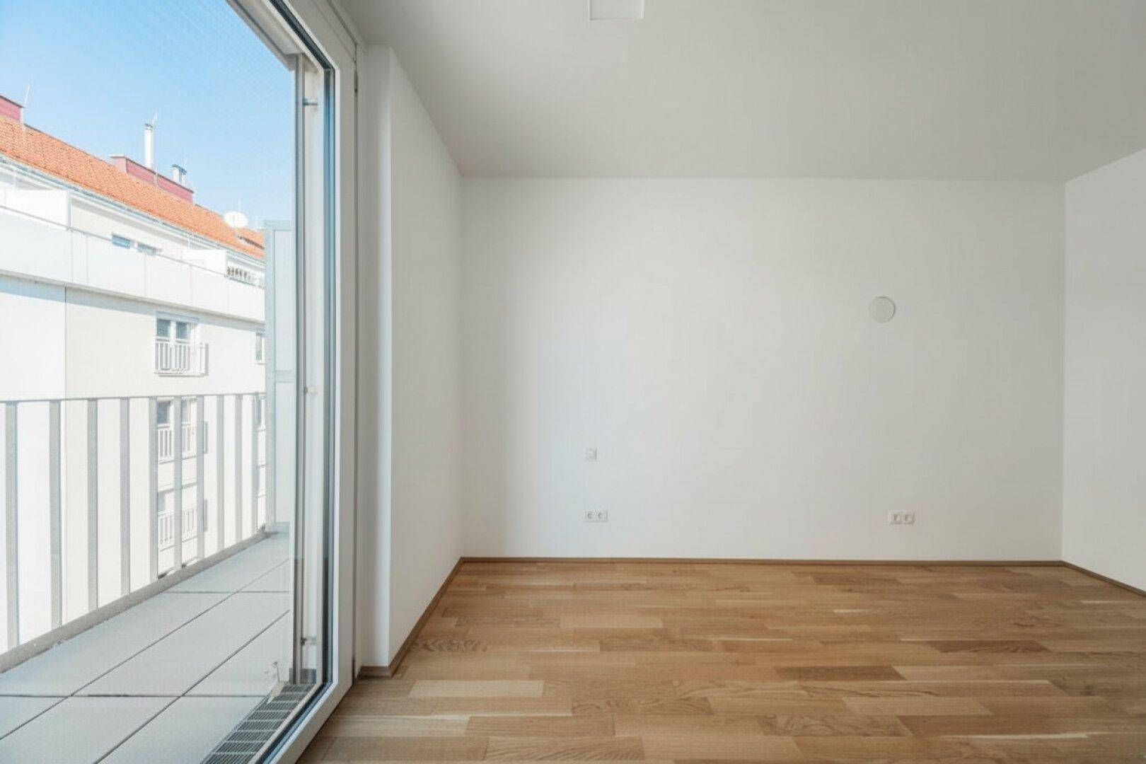 Geräumiges, leeres Zimmer mit Parkettboden und direktem Zugang zu einem sonnigen Balkon.