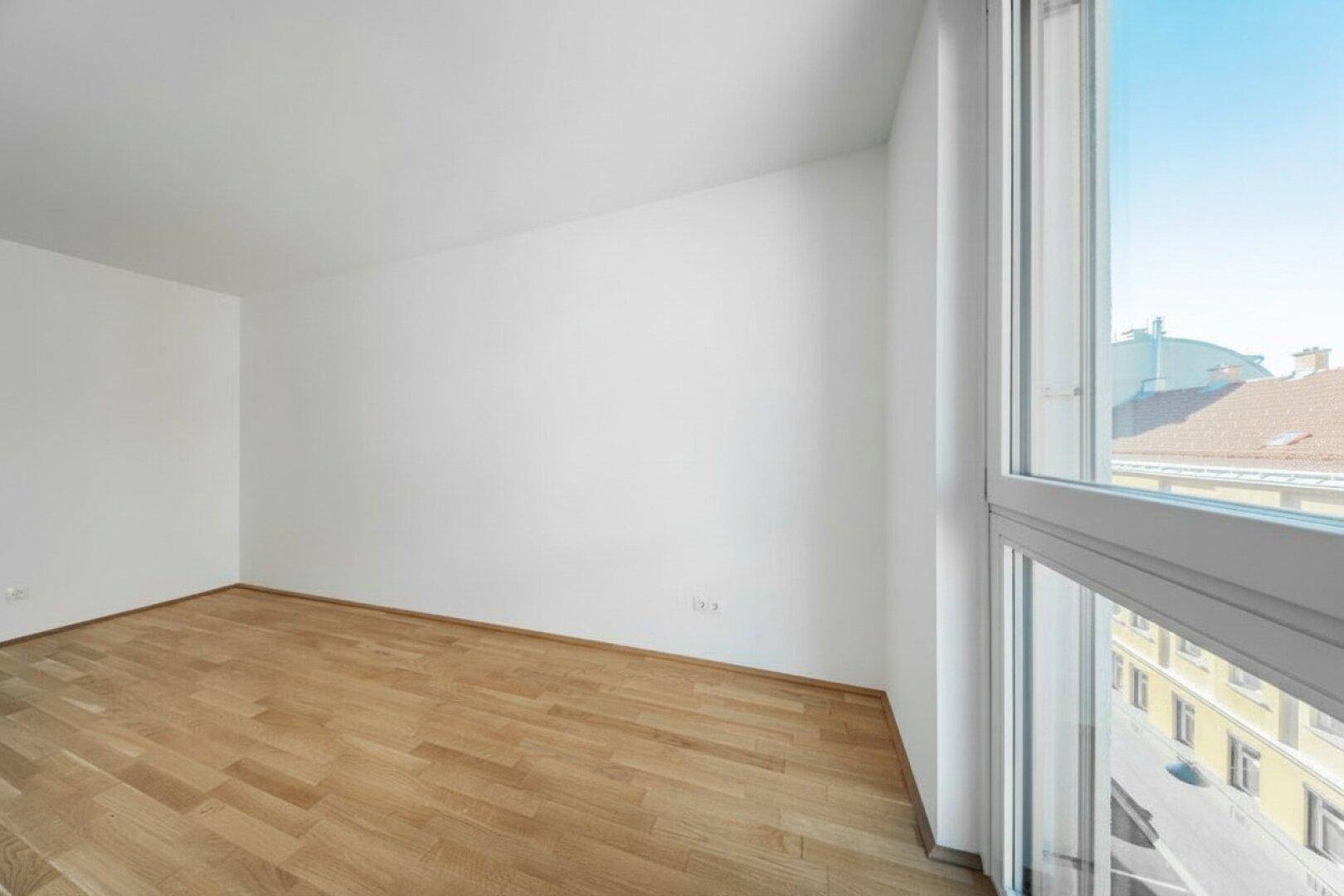 Geräumiges, helles Zimmer mit Parkettboden und einem großen Fenster, das viel Tageslicht bietet.