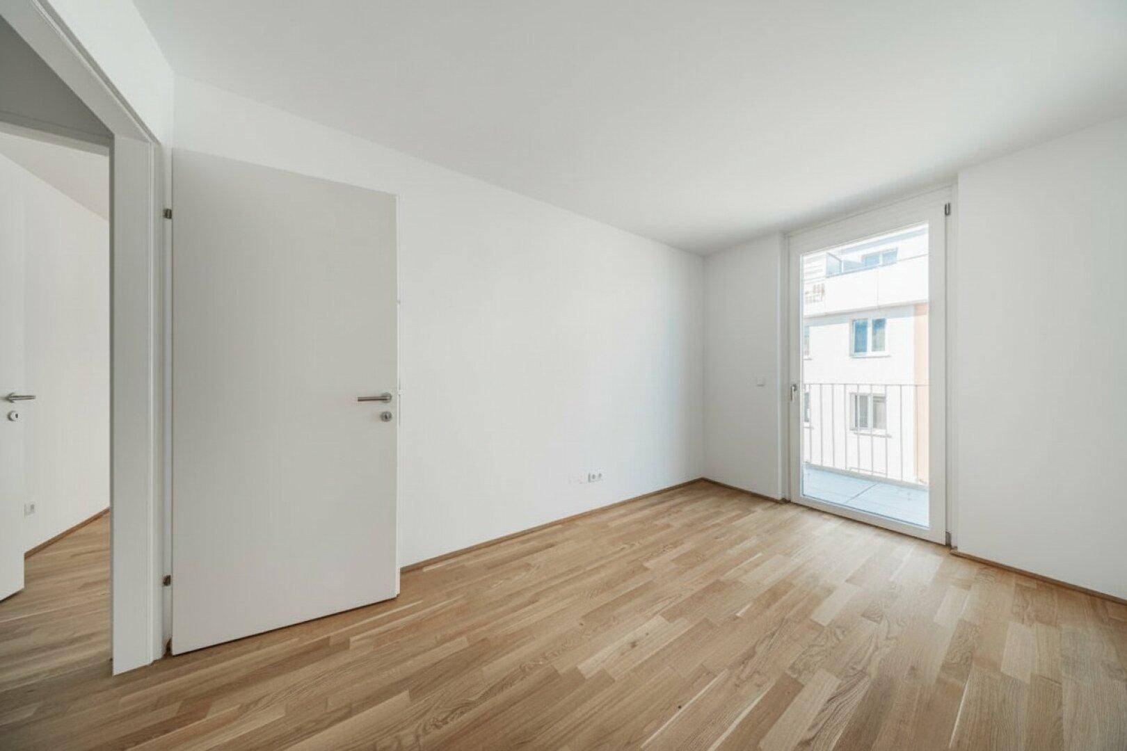 Leeres Zimmer mit Parkettboden und einem französischen Balkon, der viel Licht hereinlässt.