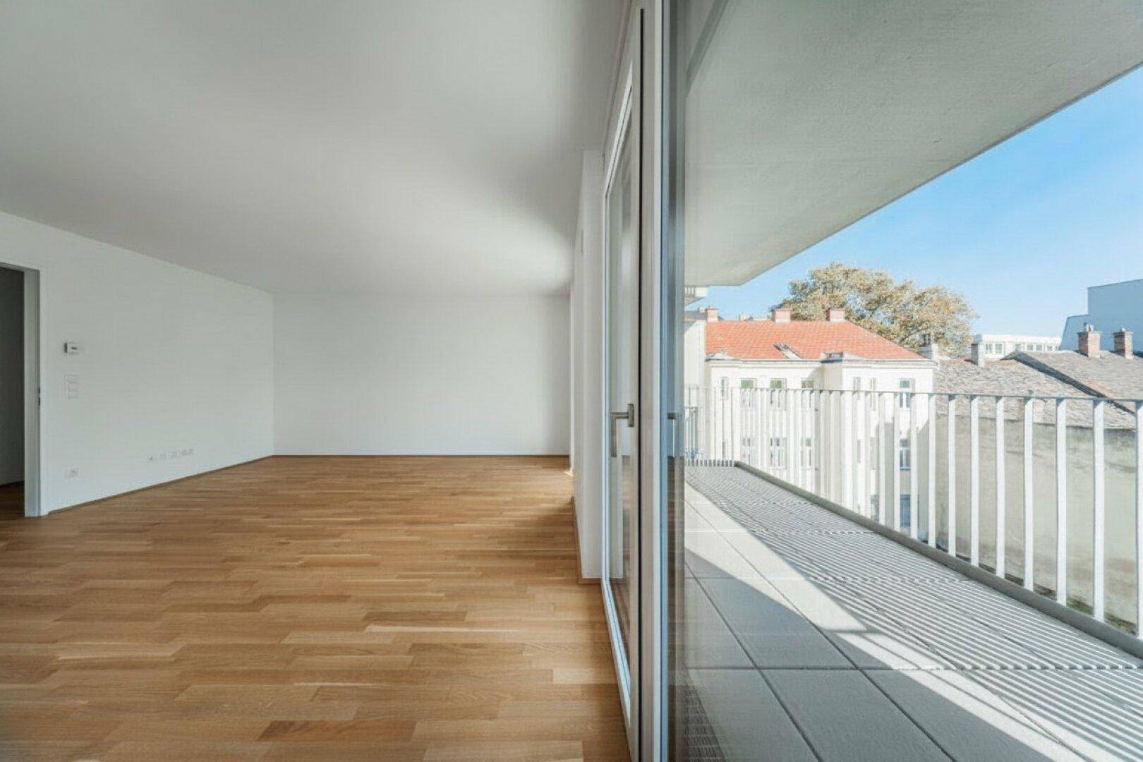 Blick aus einem hellen Wohnraum auf den Balkon mit Fliesenboden und umliegende Gebäude.