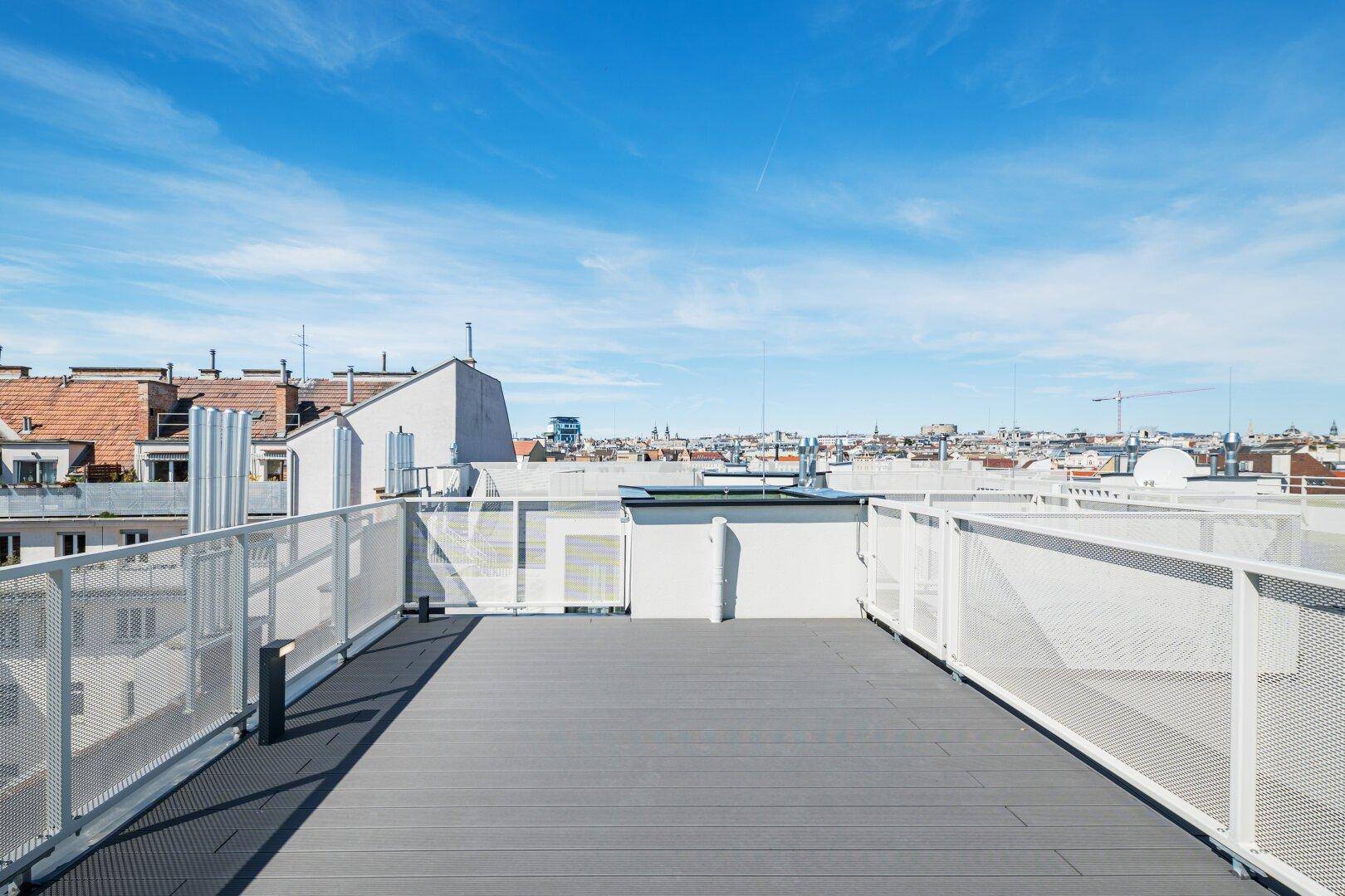 Geräumige Dachterrasse mit grauem Dielenboden und weißem Metallgeländer unter blauem Himmel.