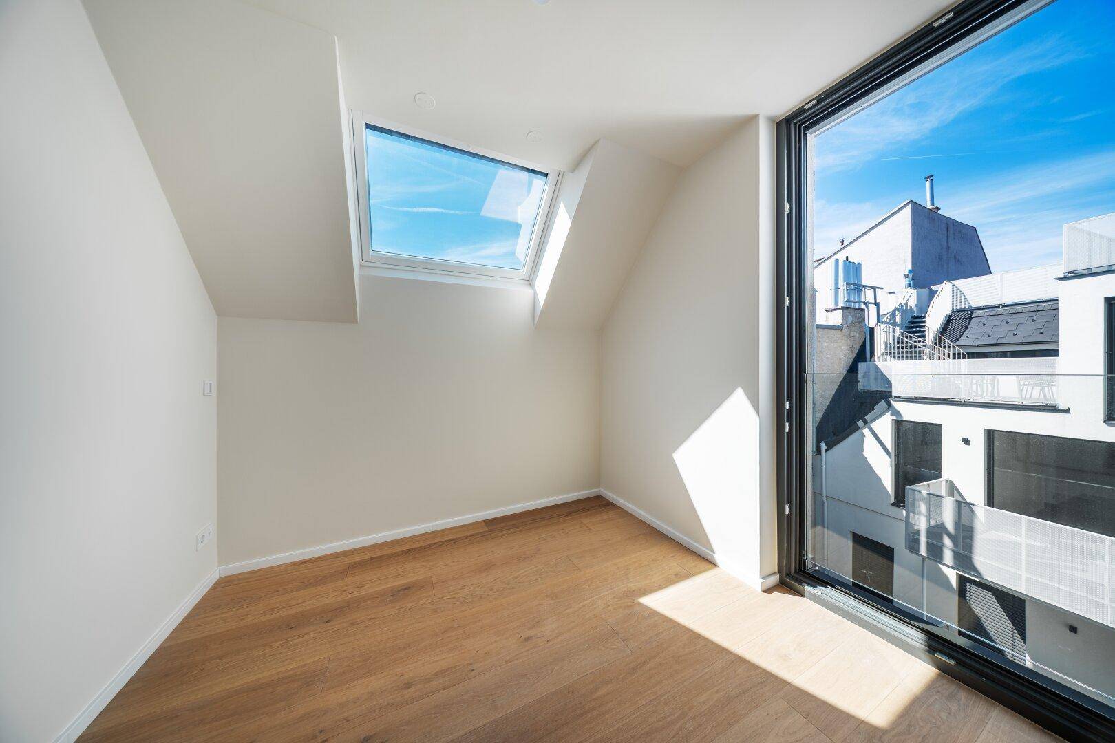 Helles Zimmer im Dachgeschoss mit Holzboden, schrägen Wänden und großem Fenster mit Stadtblick.
