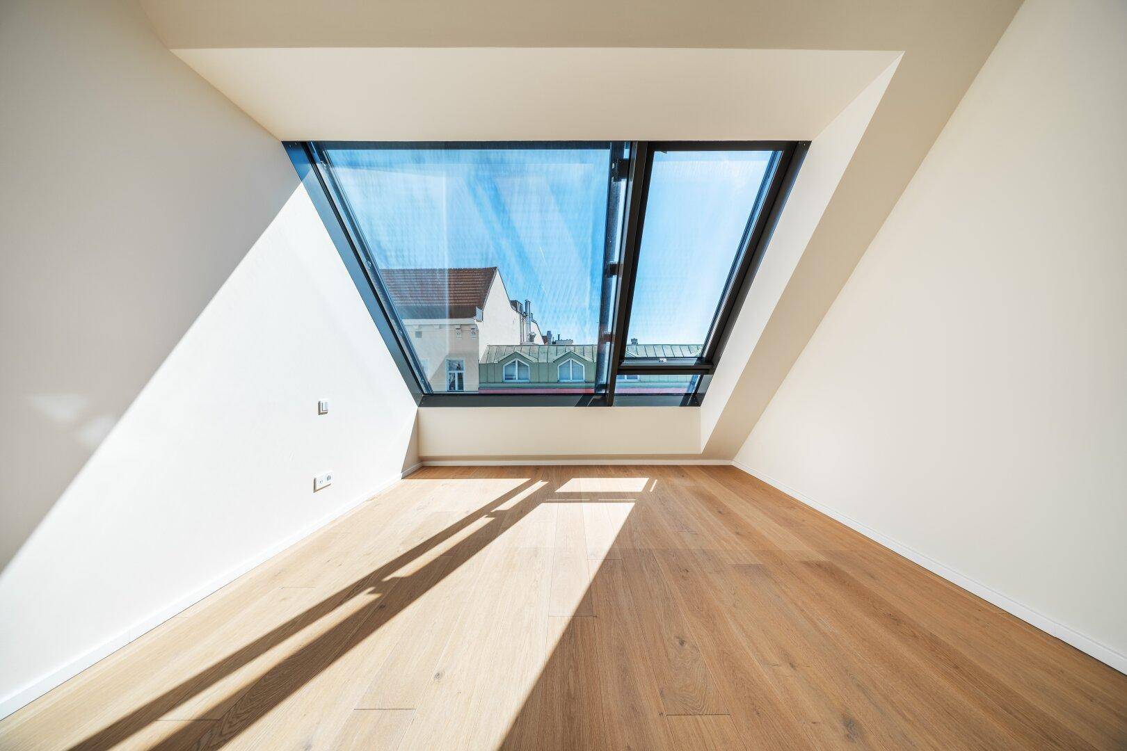 Lichtdurchflutetes Dachzimmer mit Holzboden und breiter Fensterfront, die einen weiten Blick bietet.