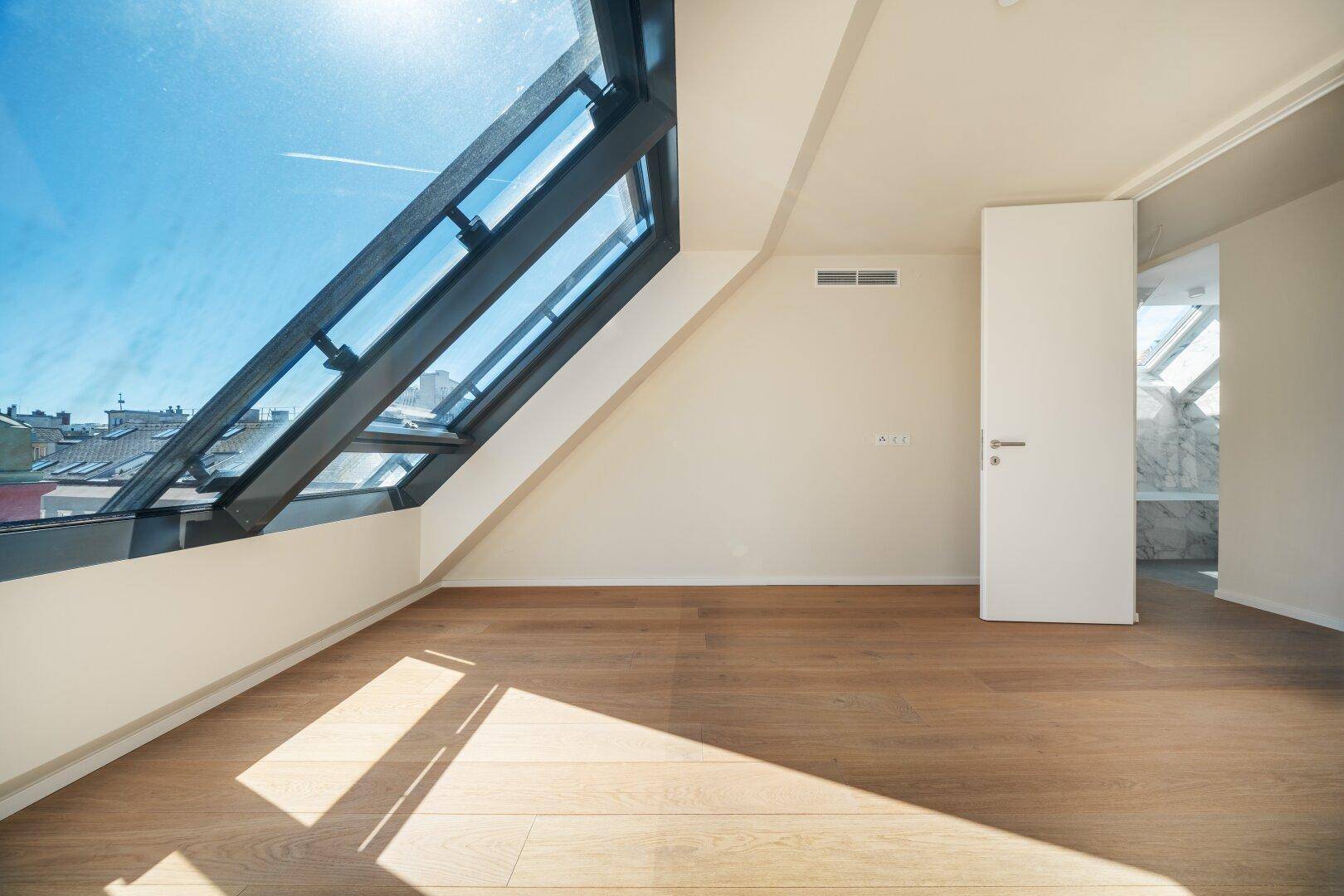Geräumiges Dachzimmer mit Holzboden, schrägen Wänden und Zugang zu einem angrenzenden Badezimmer.