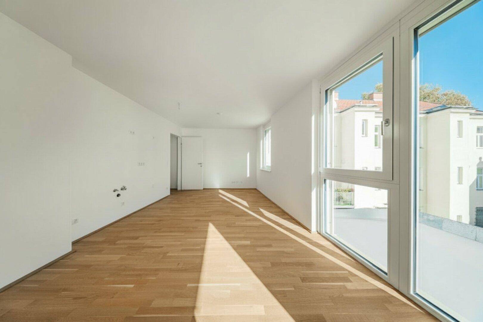 Helles, leeres Zimmer mit Parkettboden und großen Fenstern, die viel Tageslicht hereinlassen.