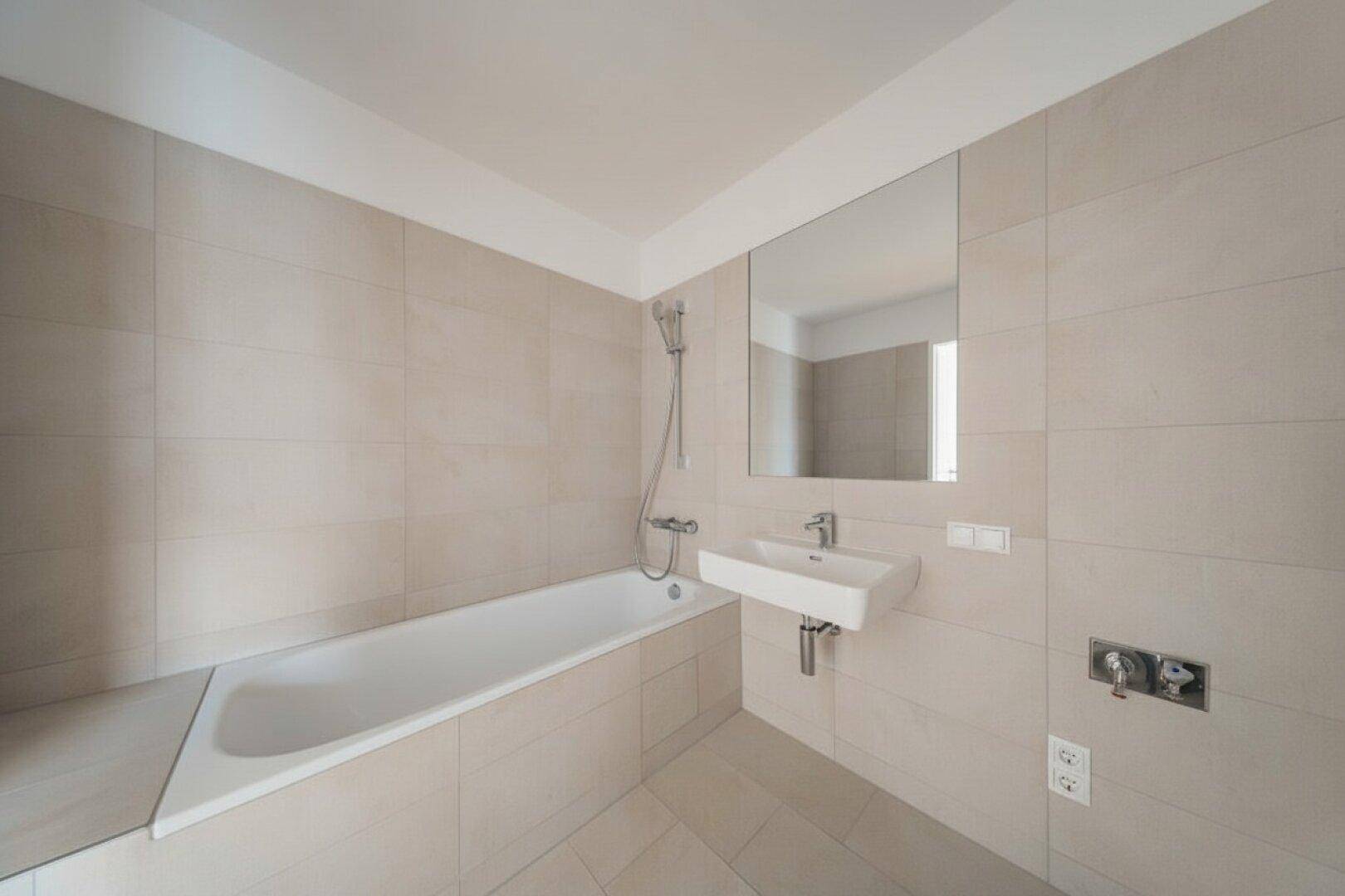 Helles Badezimmer mit Badewanne und Waschbecken, ausgestattet mit modernen Fliesen und einem Spiegel.
