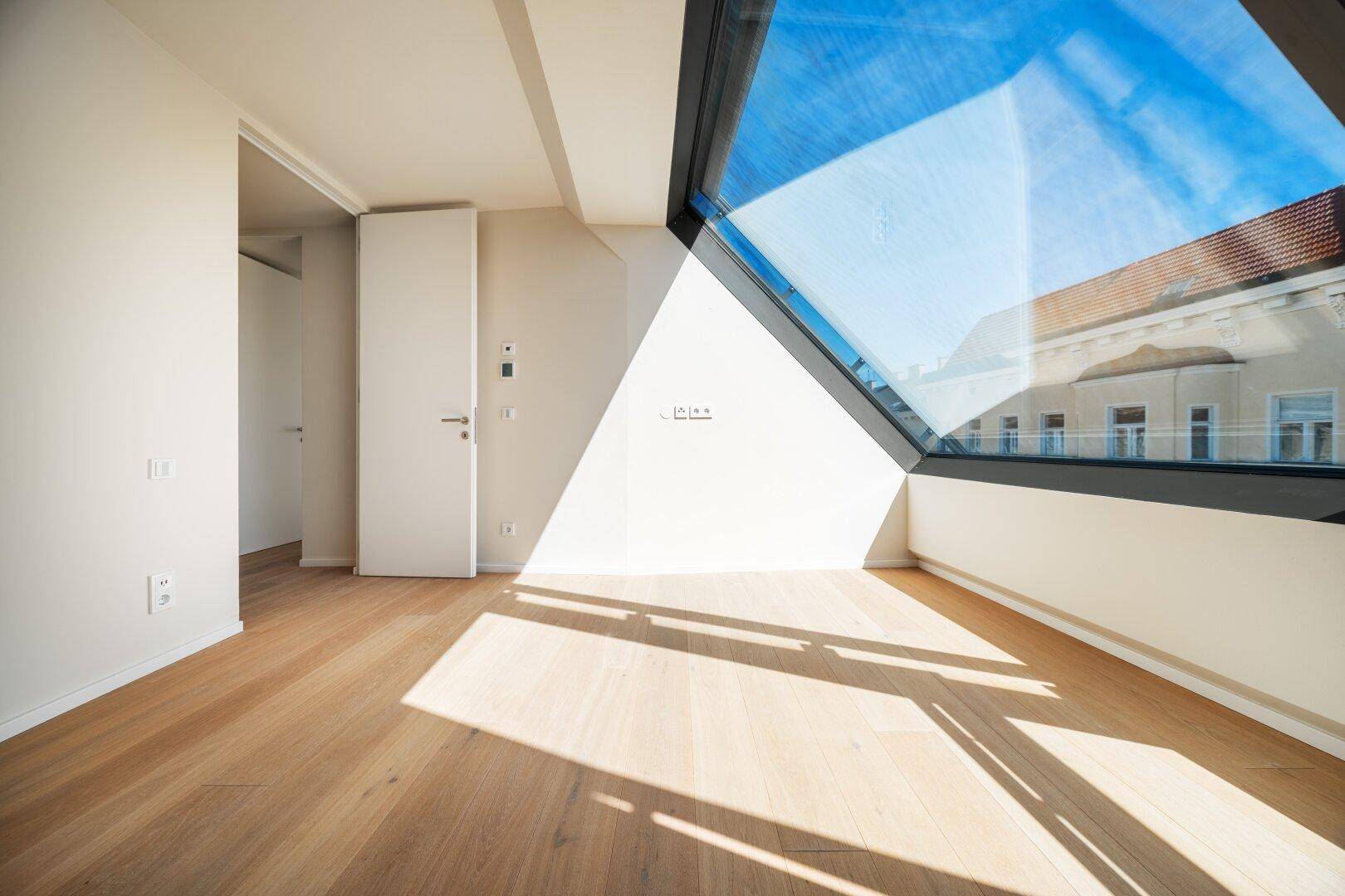 Geräumiger, heller Raum mit Holzboden und schrägem Fenster, das einen weiten Blick bietet.