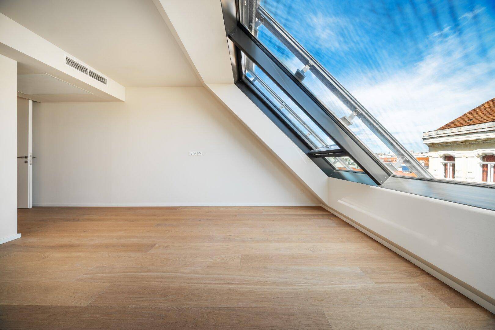Geräumiger, heller Raum mit Holzboden und großem, schrägen Fenster, das viel Licht spendet.