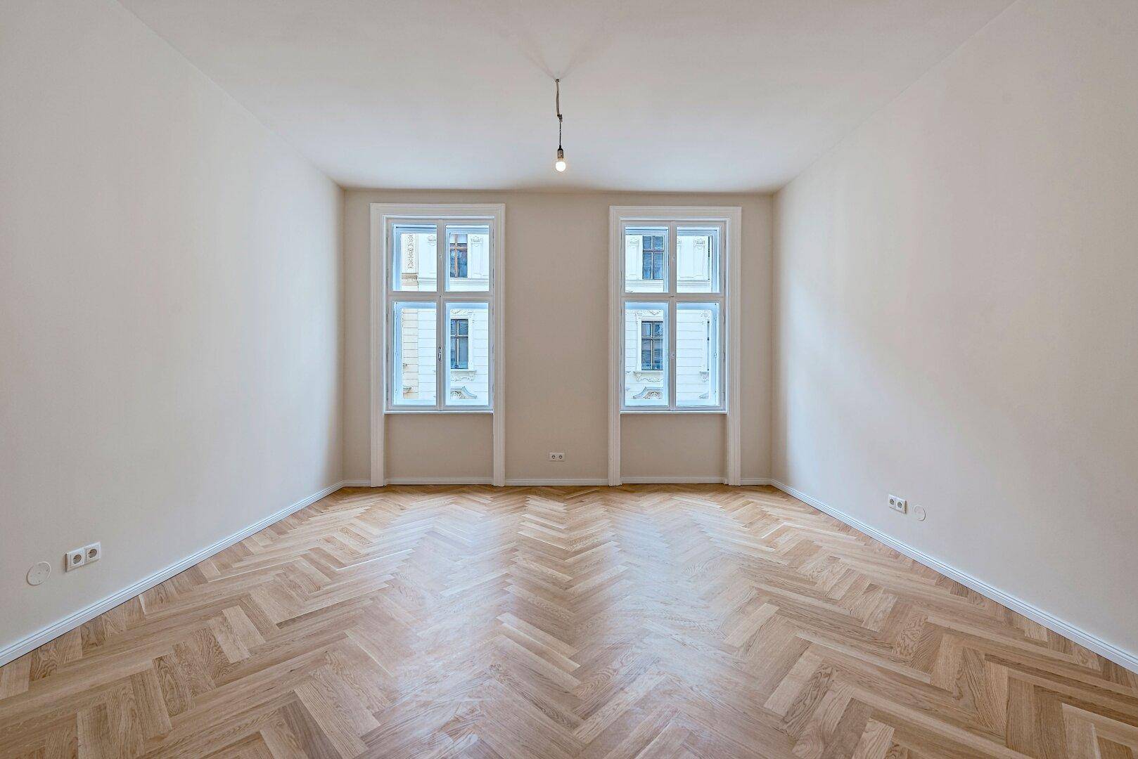 Geräumiges Zimmer mit klassischem Fischgrätparkett und zwei großen Fenstern.