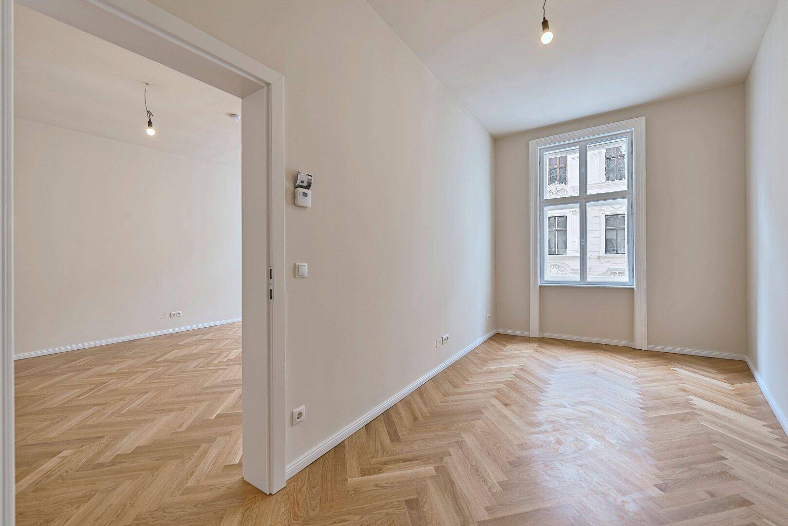 Heller Raum mit Fischgrätparkett und Zugang zu einem weiteren Zimmer.