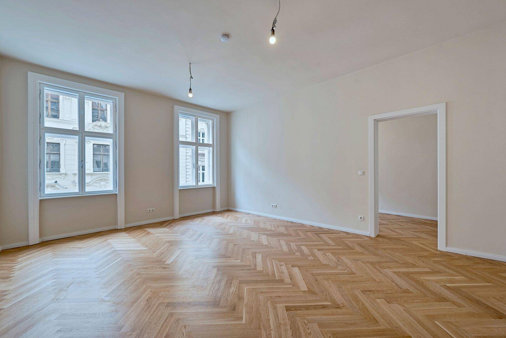 Lichtdurchflutetes Zimmer mit Fischgrätparkett und hohen Fenstern, ideal für Wohnzwecke.