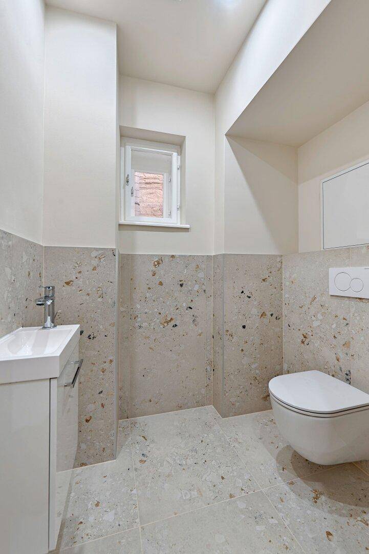 Separates WC mit wandhängender Toilette, kleinem Waschbecken und Fenster.