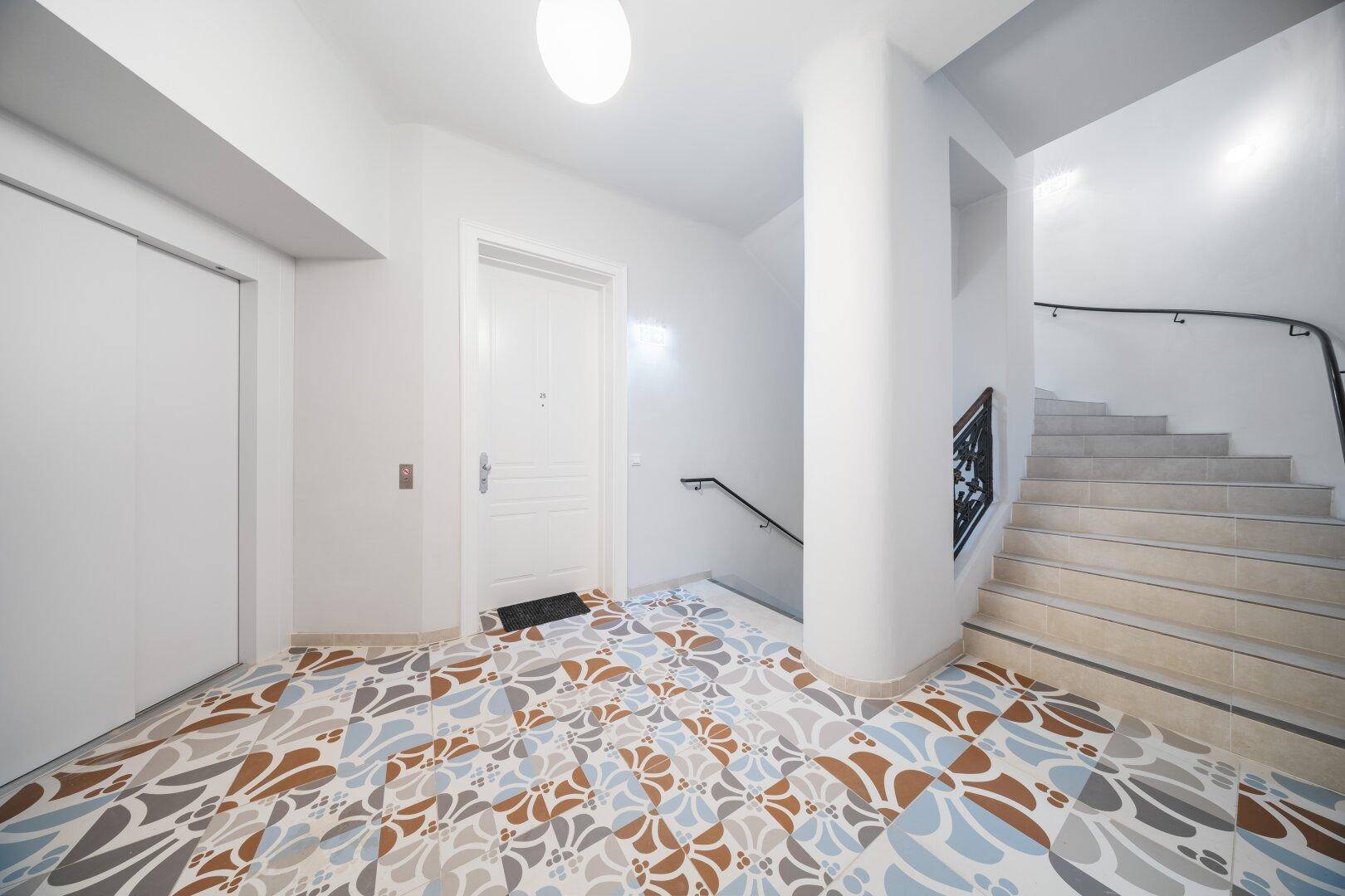 Heller Eingangsbereich mit Treppe, modernem Aufzug und dekorativem Fliesenboden.