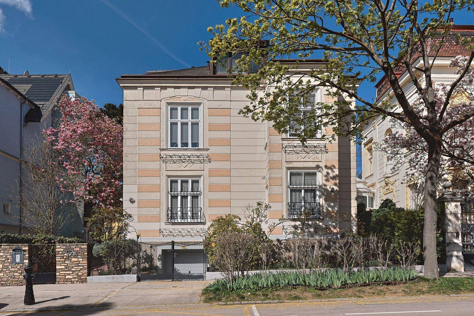 Elegante Fassade eines historischen Gebäudes mit großen Fenstern und gepflegter Vorgartenbepflanzung an der Straße.