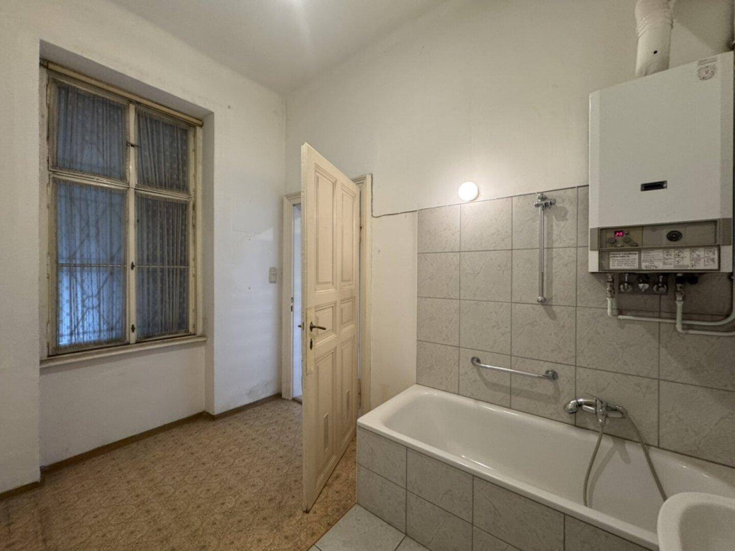 Badezimmer mit Badewanne, kleinem Waschbecken und großem Fenster, das Renovierungsbedarf zeigt.